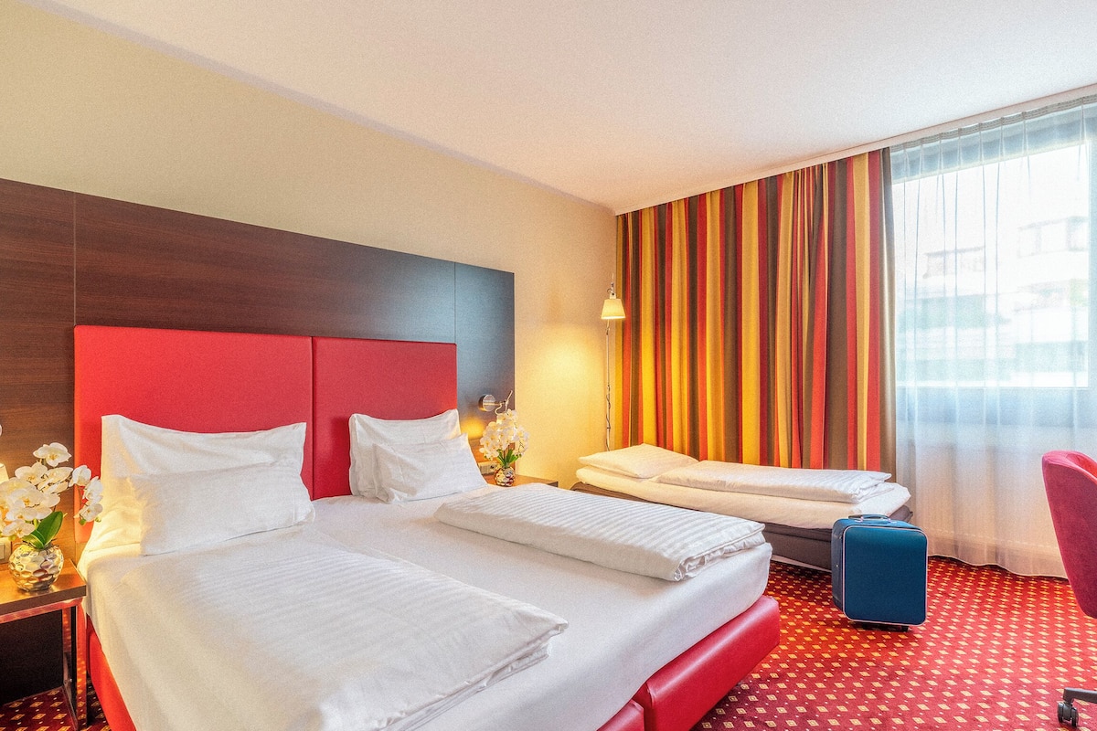 Una habitación de hotel con una cama grande, una cama más pequeña, un escritorio con lámpara y una silla.