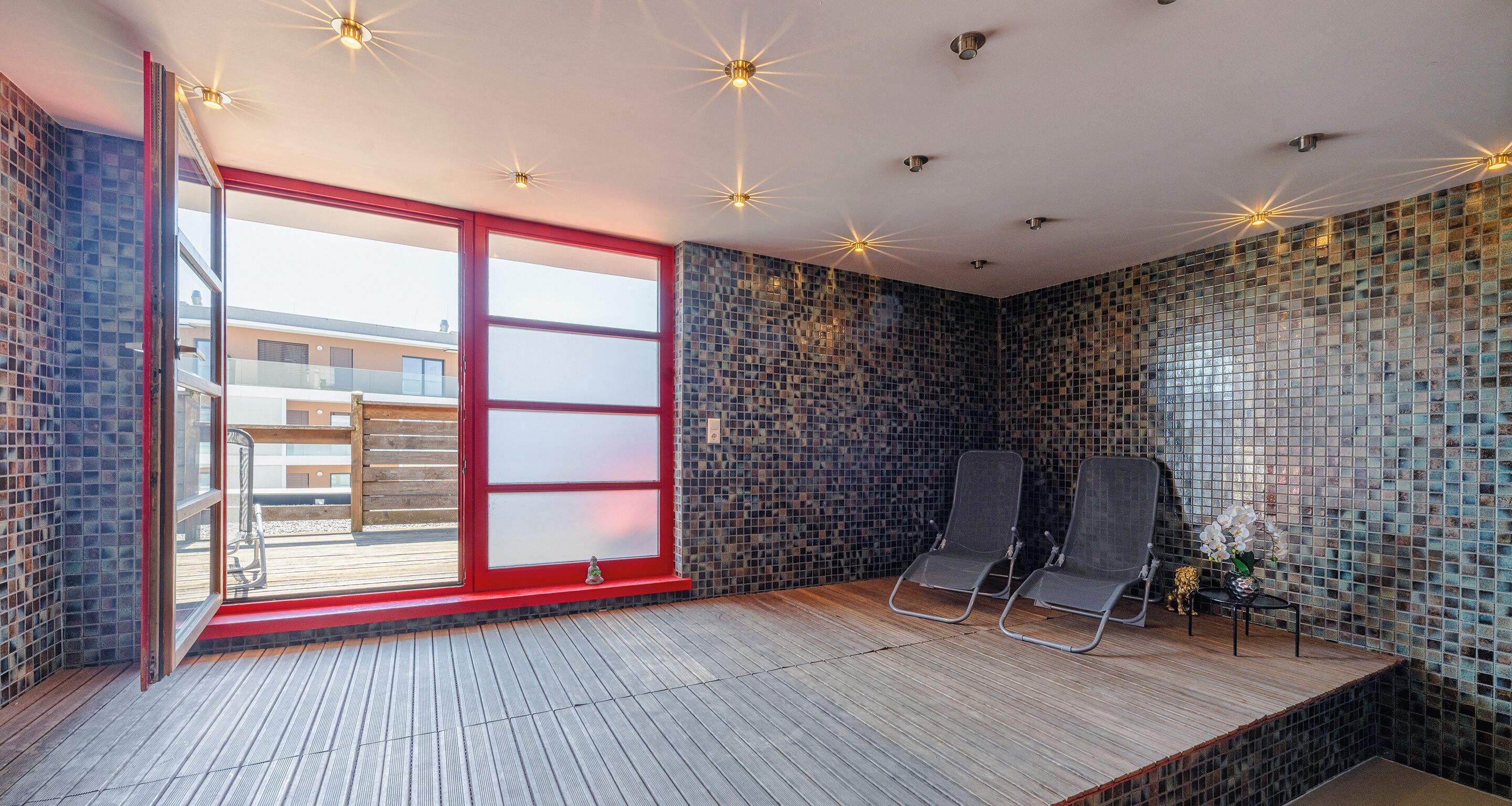 A spa room with a wooden floor, two black lounge chairs, a small table with a vase of flowers, and a red-framed glass door leading to an outdoor area.