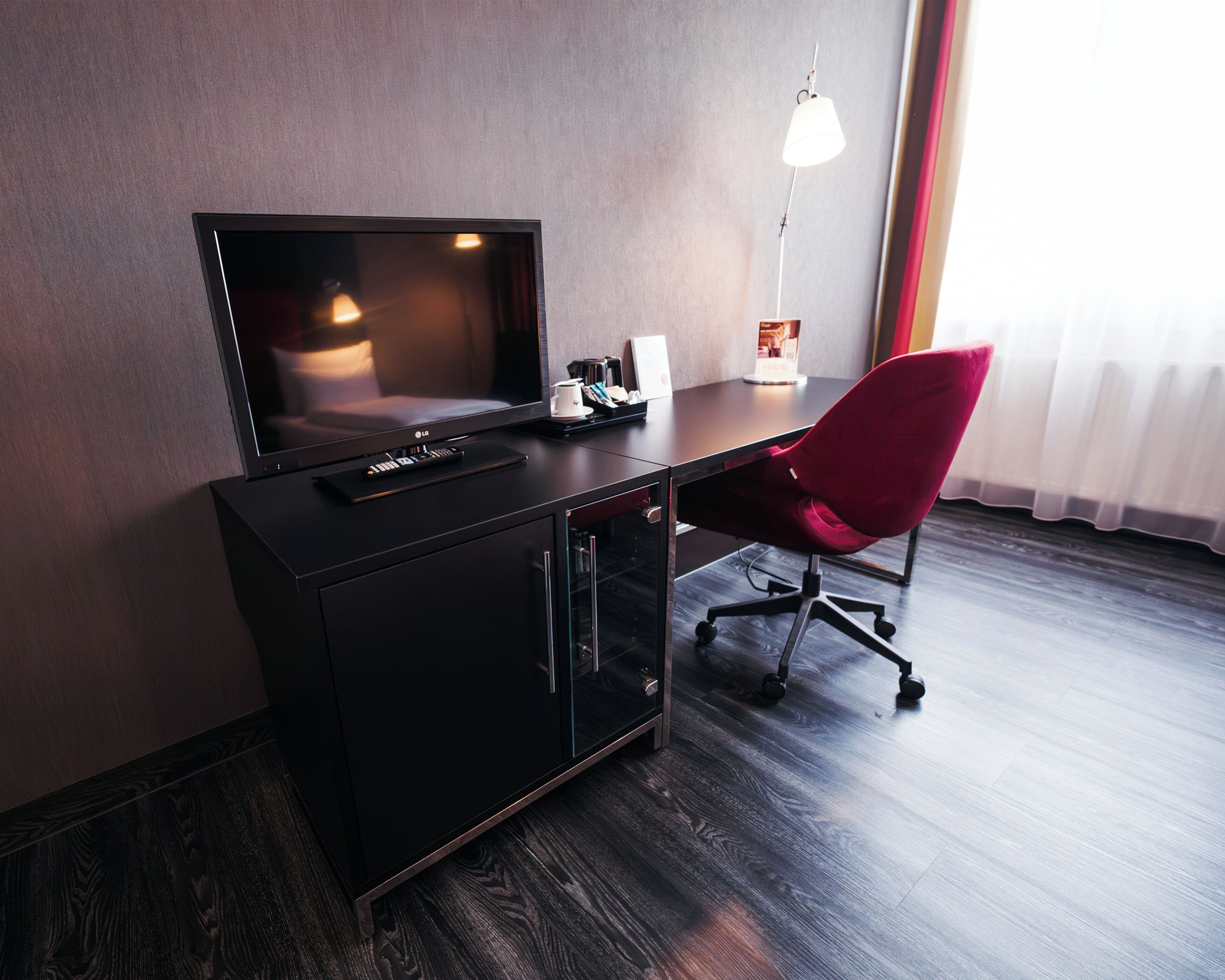 A modern hotel room with a flat-screen TV, a red office chair, a desk with a lamp, and a large window with sheer curtains.