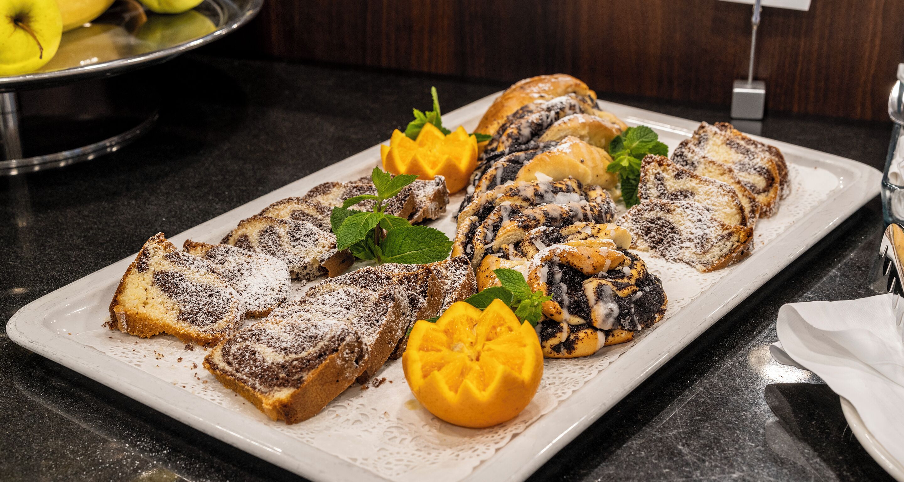 Une assiette de petit-déjeuner comprenant des petits pains, une part de gâteau et une demi-mangue, le tout présenté sur une assiette blanche.