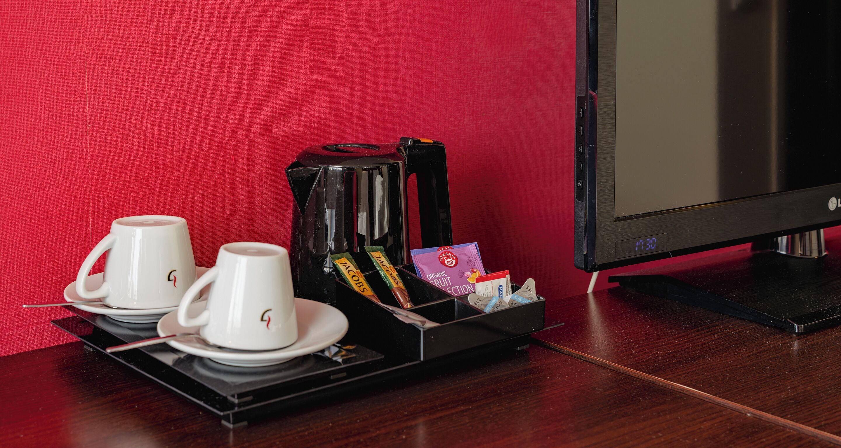 A coffee maker, two white cups, and a tray with snacks and a card on a wooden table against a red wall.