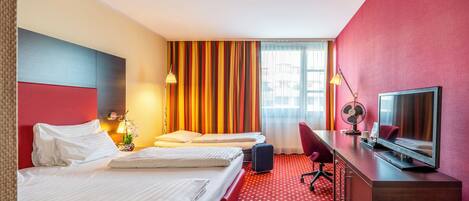 Habitación de hotel con dos camas, un escritorio con televisor, lámpara de escritorio, ventilador y ventana con cortinas.