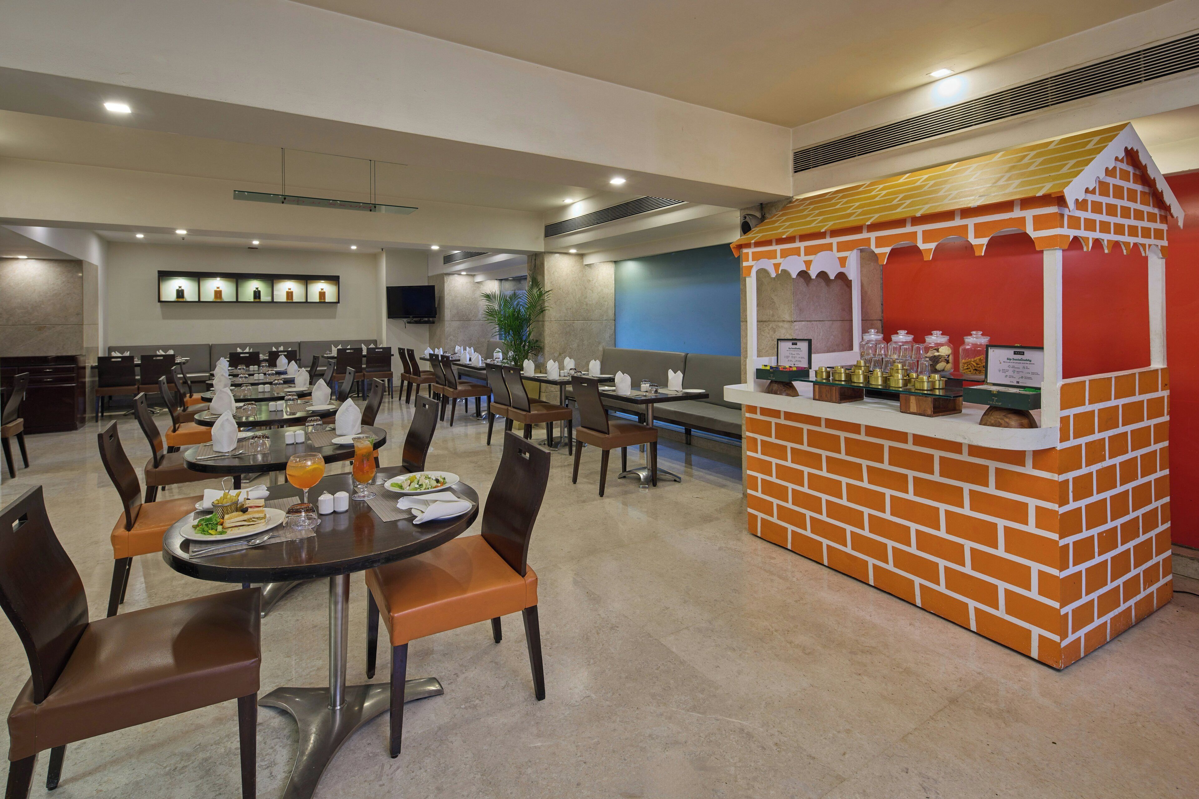 A hotel dining area with multiple tables and chairs, a brick kiosk, and a flat-screen TV mounted on the wall.