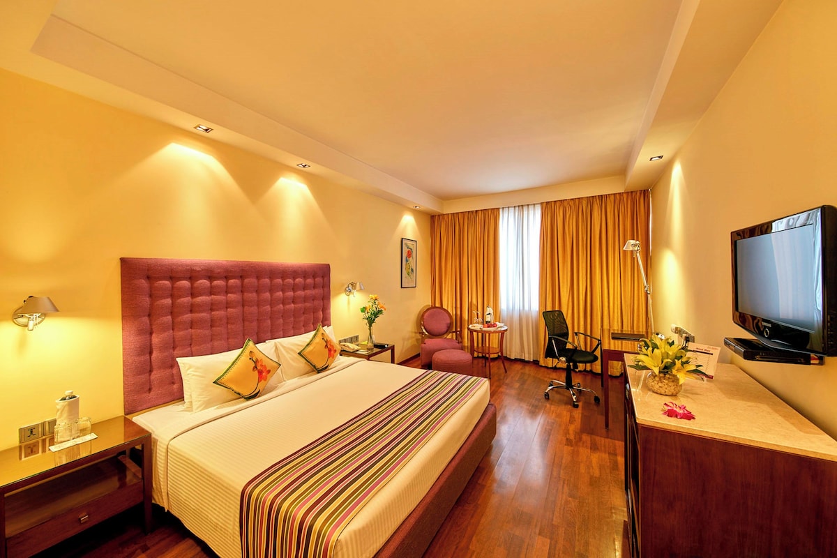 A hotel room with a large bed, a desk with a television, a chair, and a small table with flowers.