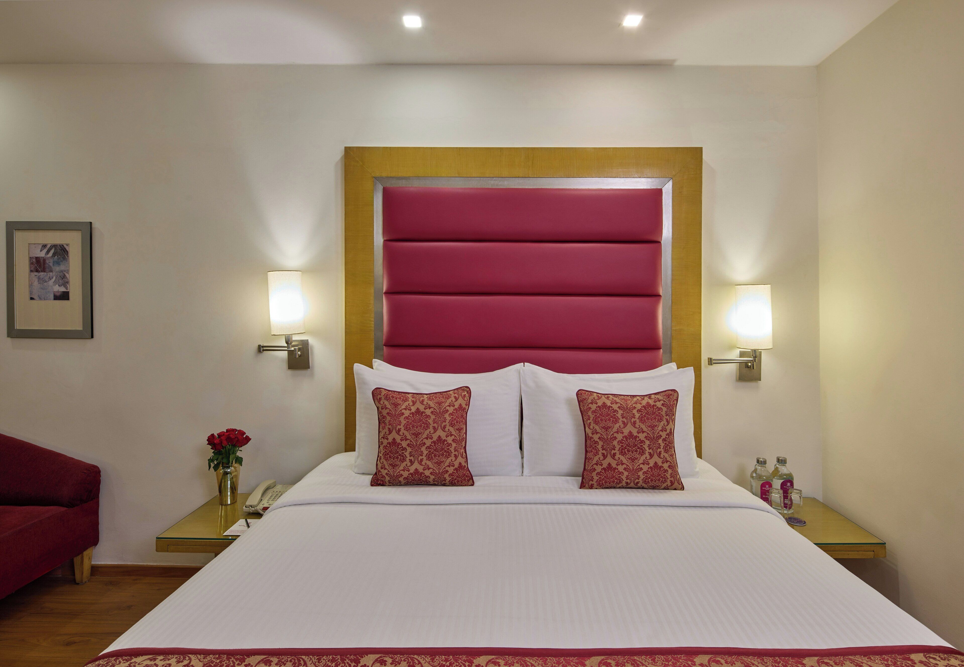 Une chambre d’hôtel avec un grand lit, deux lampes de chevet, une table de chevet avec un téléphone et un vase de fleurs, et un fauteuil rouge.