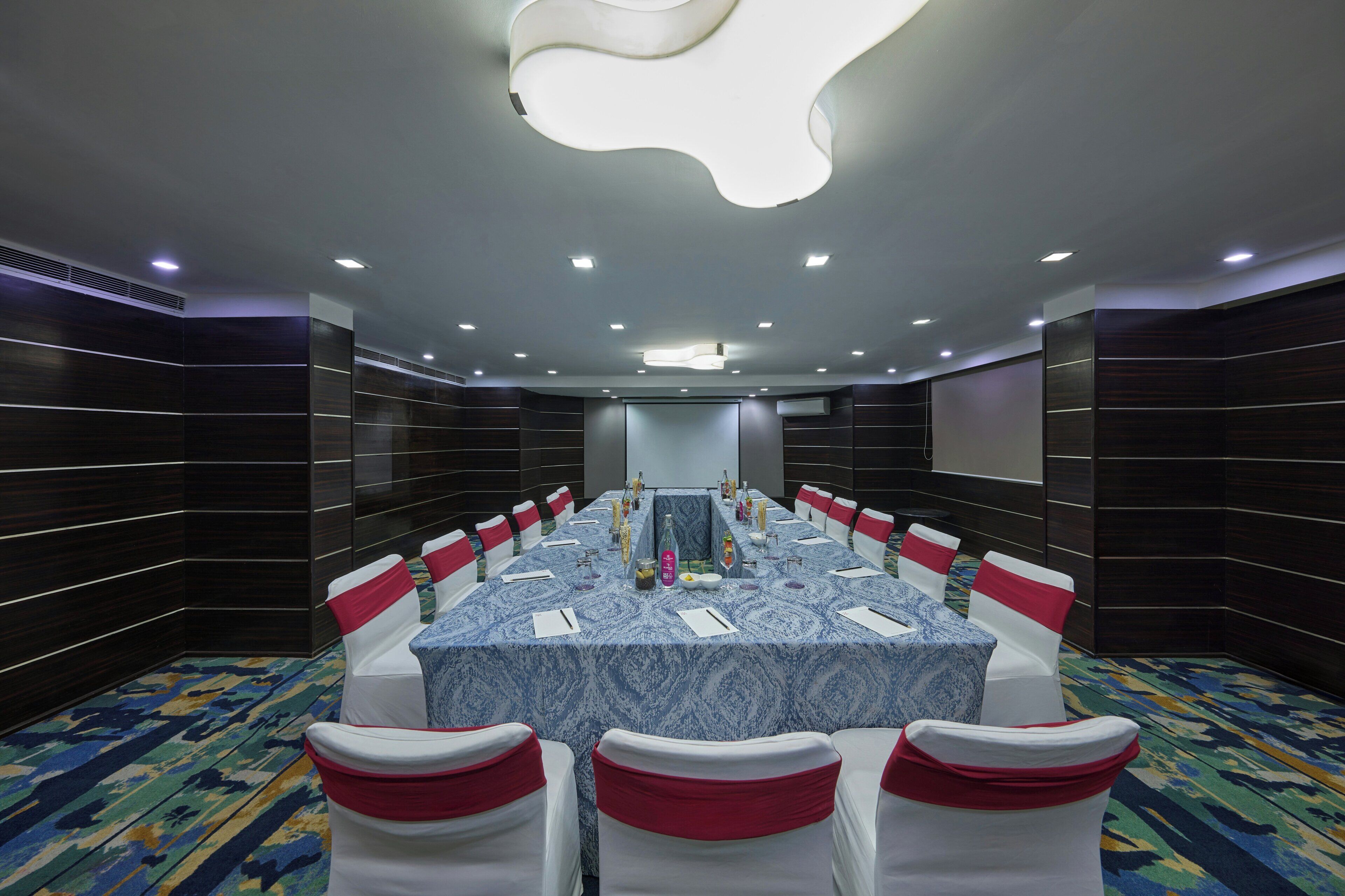 Un salón de conferencias con una larga mesa, sillas blancas con cintas rojas y una alfombra estampada.