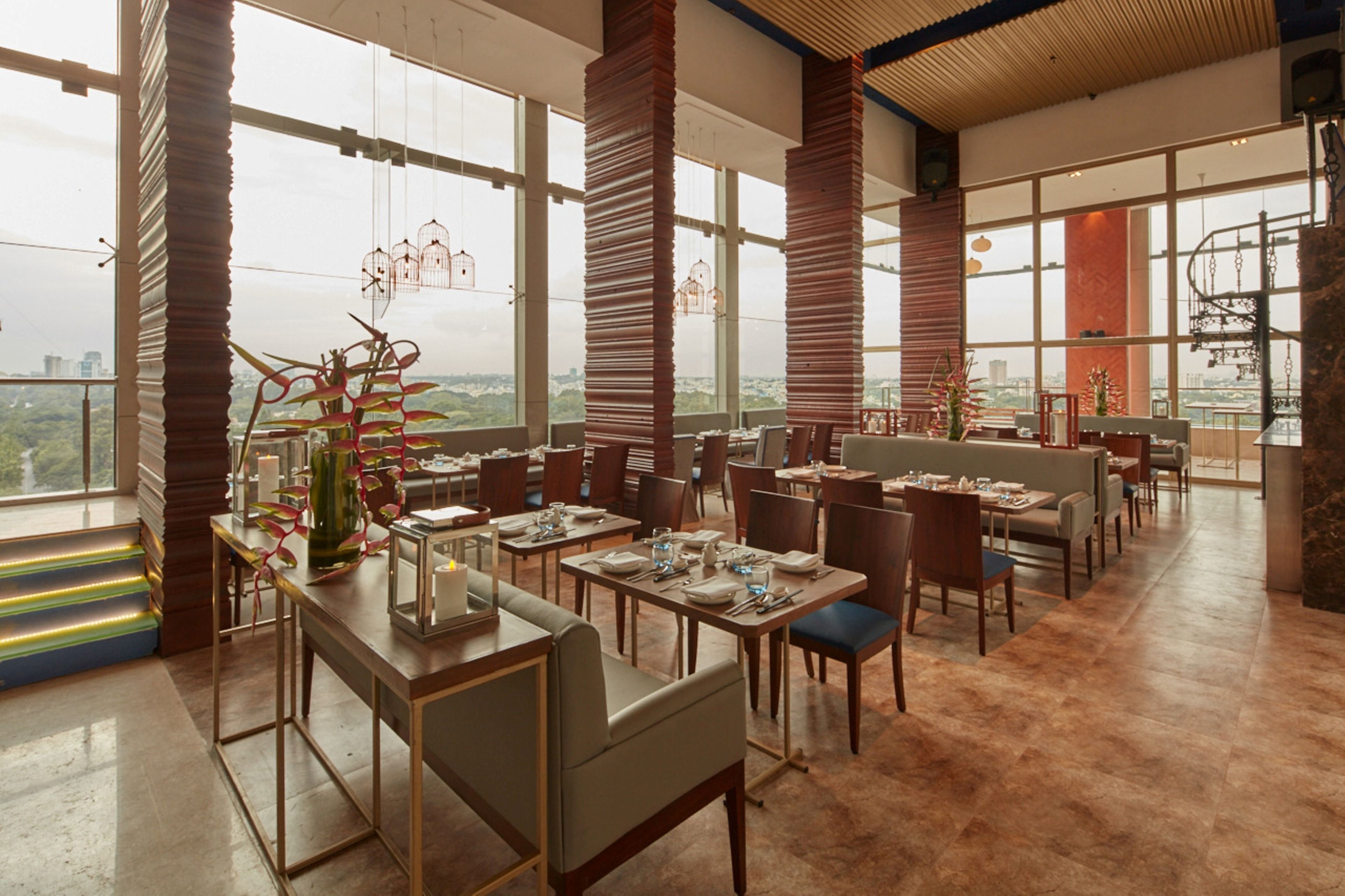 Un restaurante con techos altos, ventanas amplias y vistas de la ciudad. Las mesas están dispuestas con platos, vasos y cubiertos.