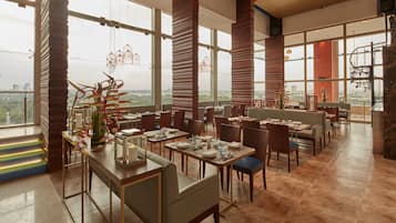 Un restaurante con techos altos, ventanas amplias y vistas de la ciudad. Las mesas están dispuestas con platos, vasos y cubiertos.
