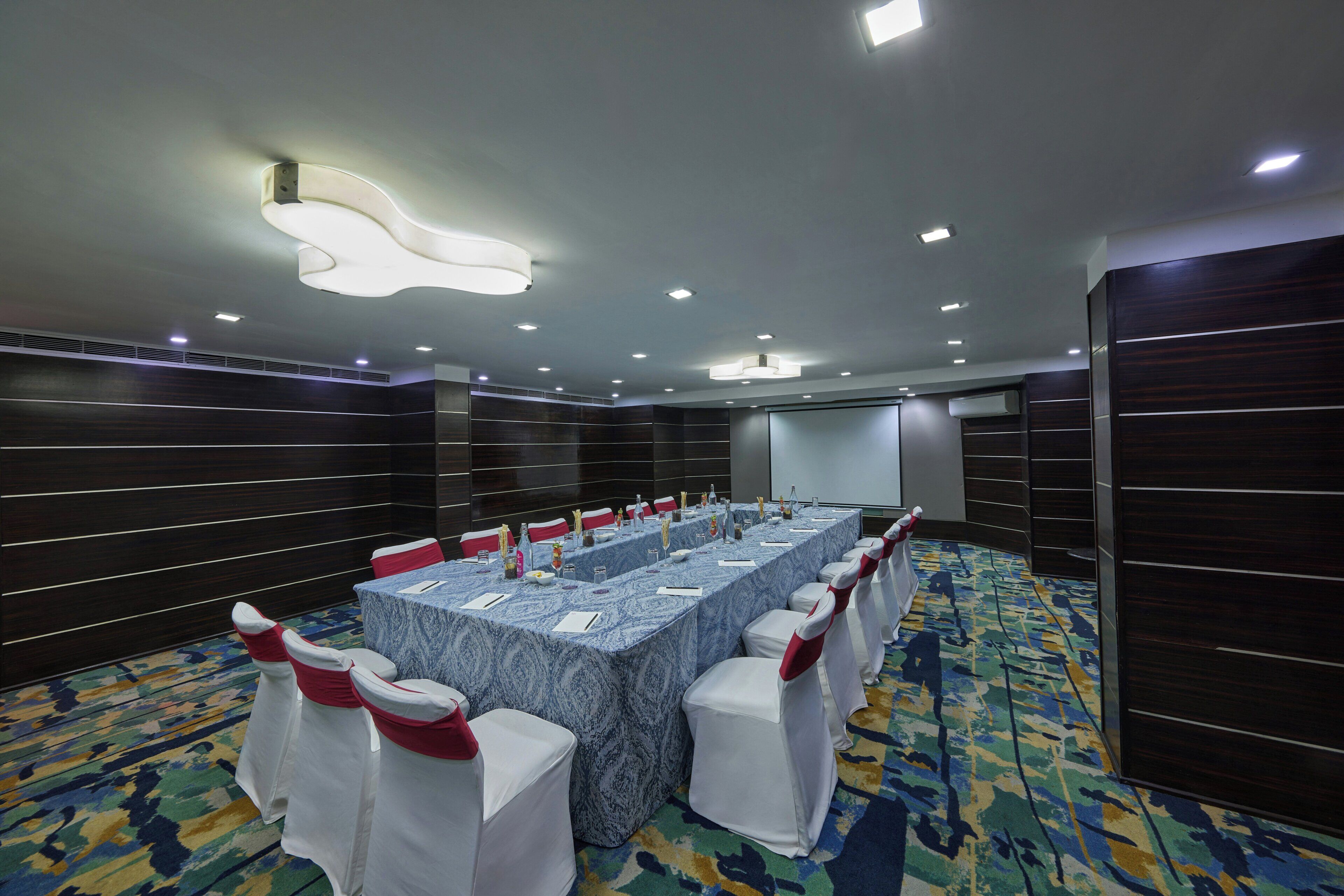 Un salón de conferencias con una larga mesa, sillas blancas adornadas con cintas rojas y una pantalla de proyector.