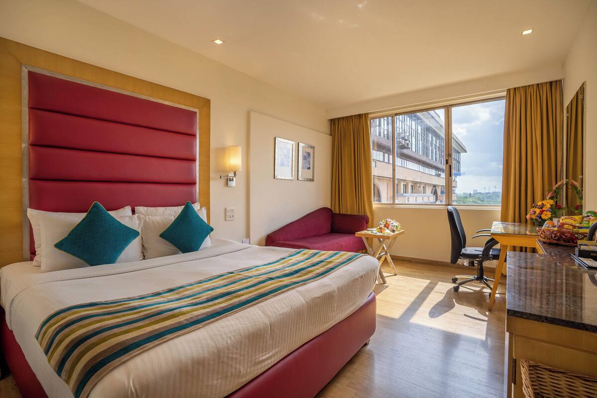 Une chambre d’hôtel avec un grand lit, une tête de lit rouge, un bureau avec une chaise, une petite table avec une lampe, et une vue sur un immeuble par la fenêtre.