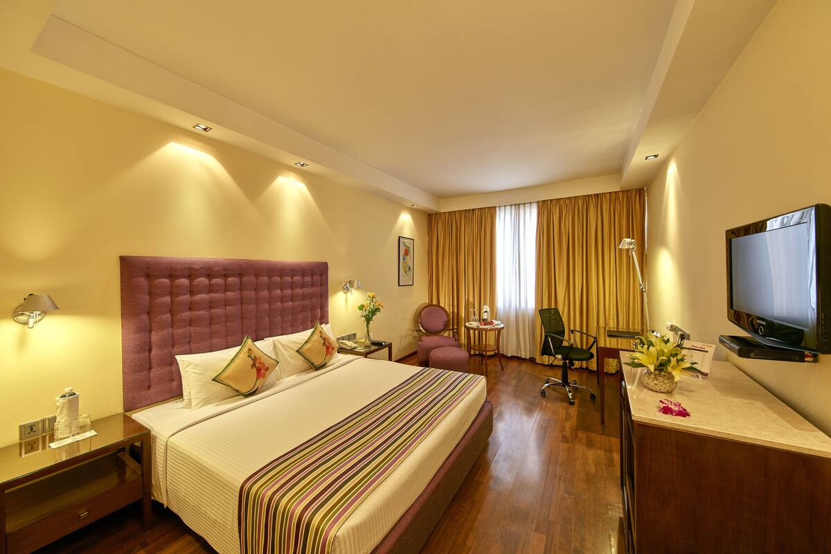 Une chambre d’hôtel avec un grand lit, un bureau avec une télévision, une chaise, une lampe de bureau et une petite table sur laquelle est posé un vase de fleurs.