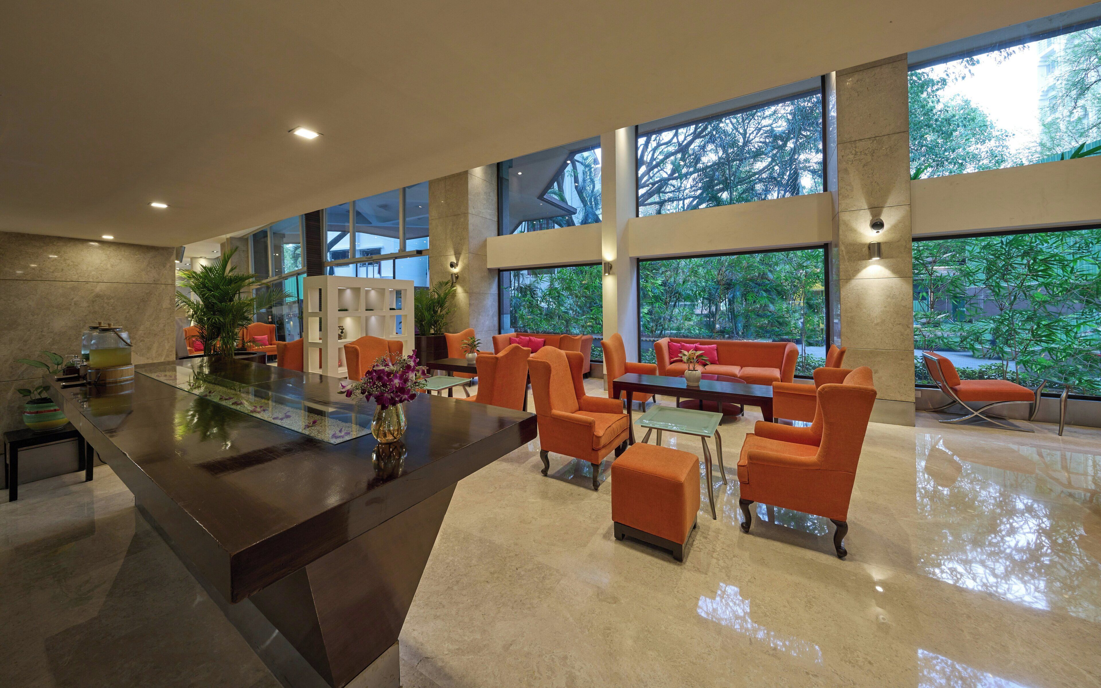 A spacious lobby with a central reception desk, seating area with orange chairs, large windows offering views of greenery, and a modern interior design.