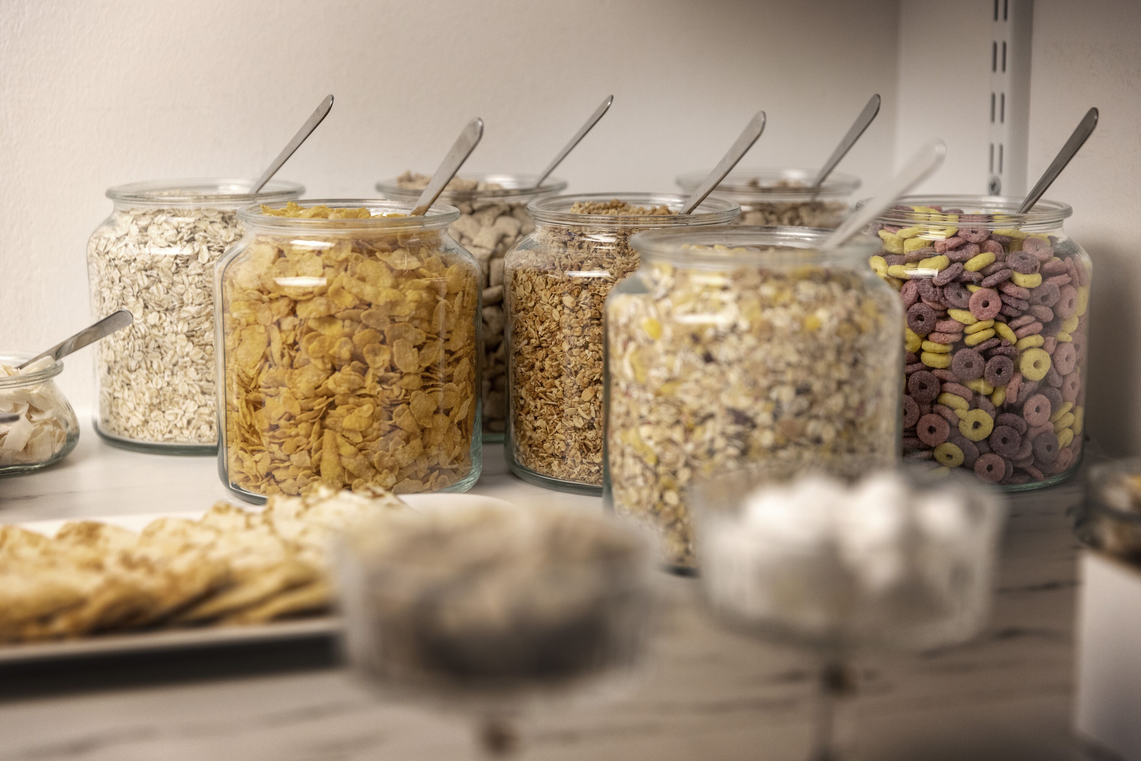 Variedad de alimentos en frascos de vidrio, con cucharas, incluyendo cereales y aperitivos, sobre una mesa.