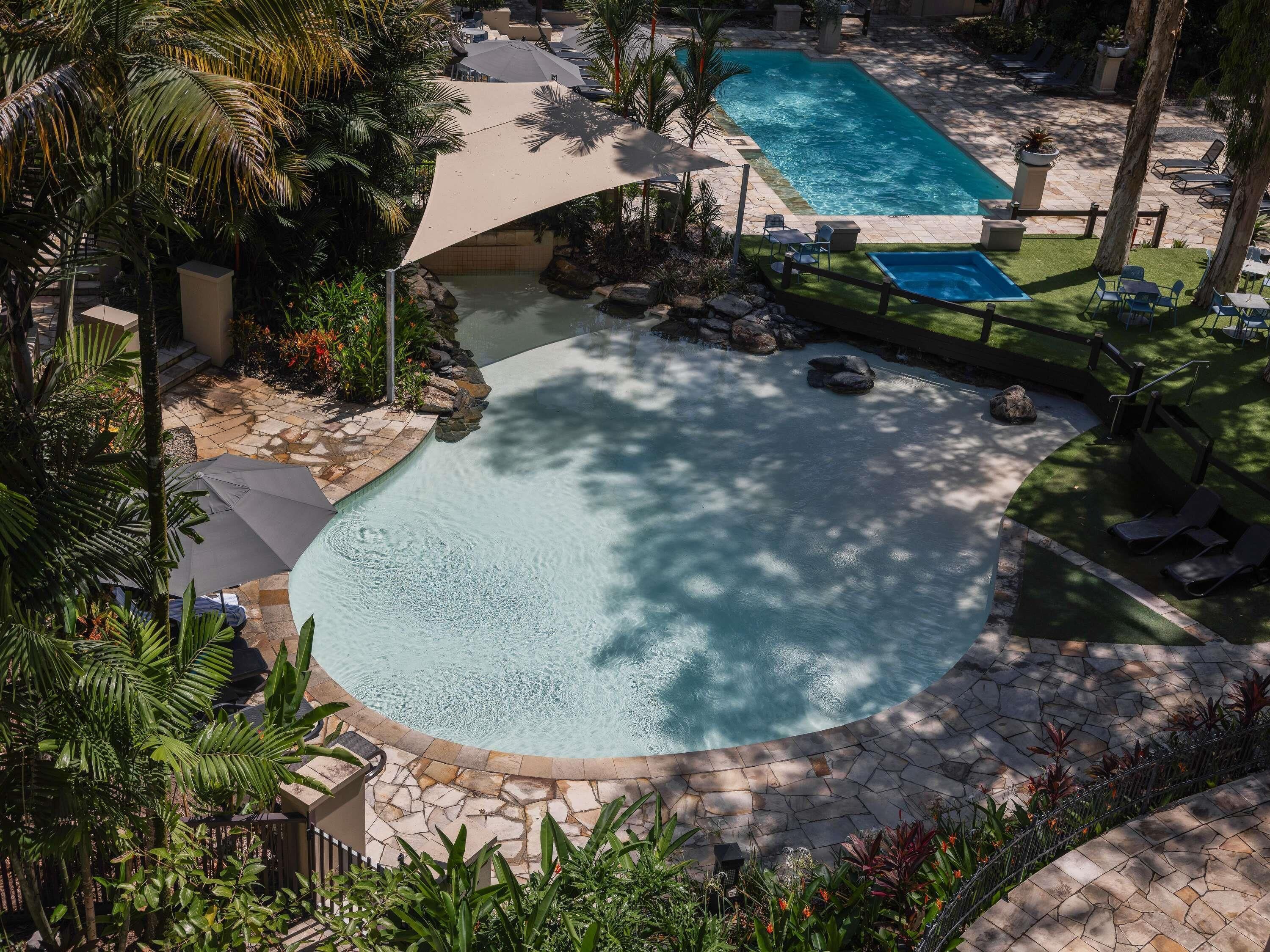 A large outdoor swimming pool with a stone border, surrounded by palm trees and lounge chairs.