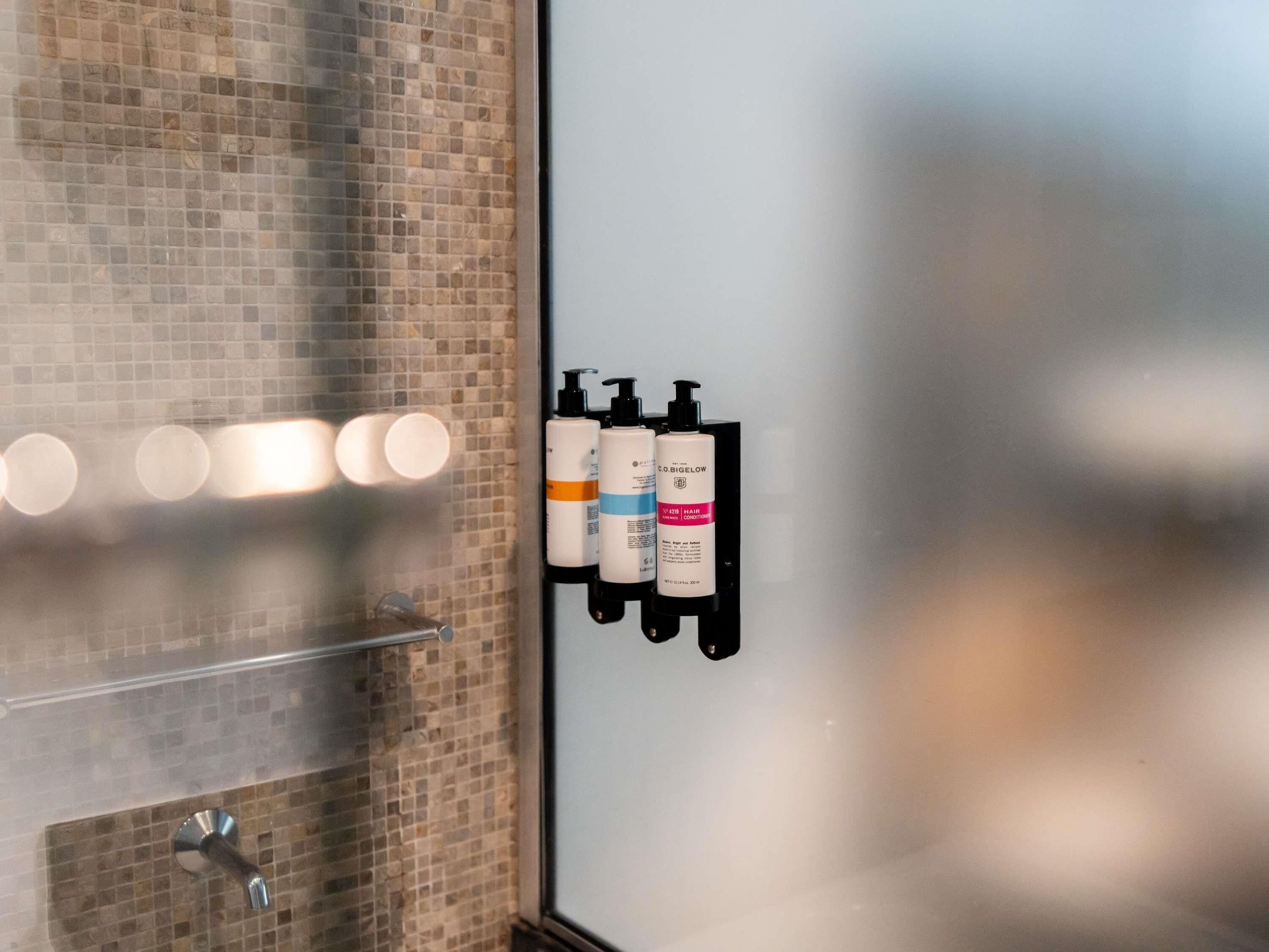 A bathroom with a tiled wall, a mirror, and two bottles of hair conditioner on a wall-mounted dispenser.