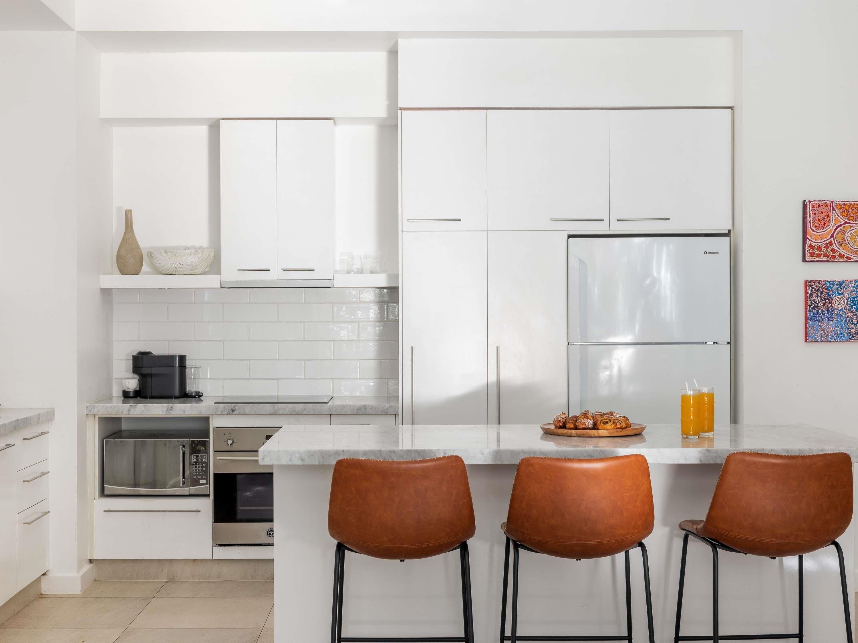 A modern kitchen with white cabinets, a stainless steel refrigerator, a coffee maker, and three brown leather bar stools.
