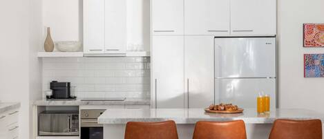 A modern kitchen with white cabinets, a stainless steel refrigerator, a coffee maker, and three brown leather bar stools.