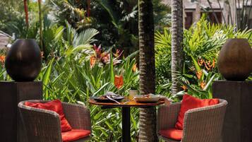 A table set for two with wicker chairs and red cushions, surrounded by lush tropical plants.