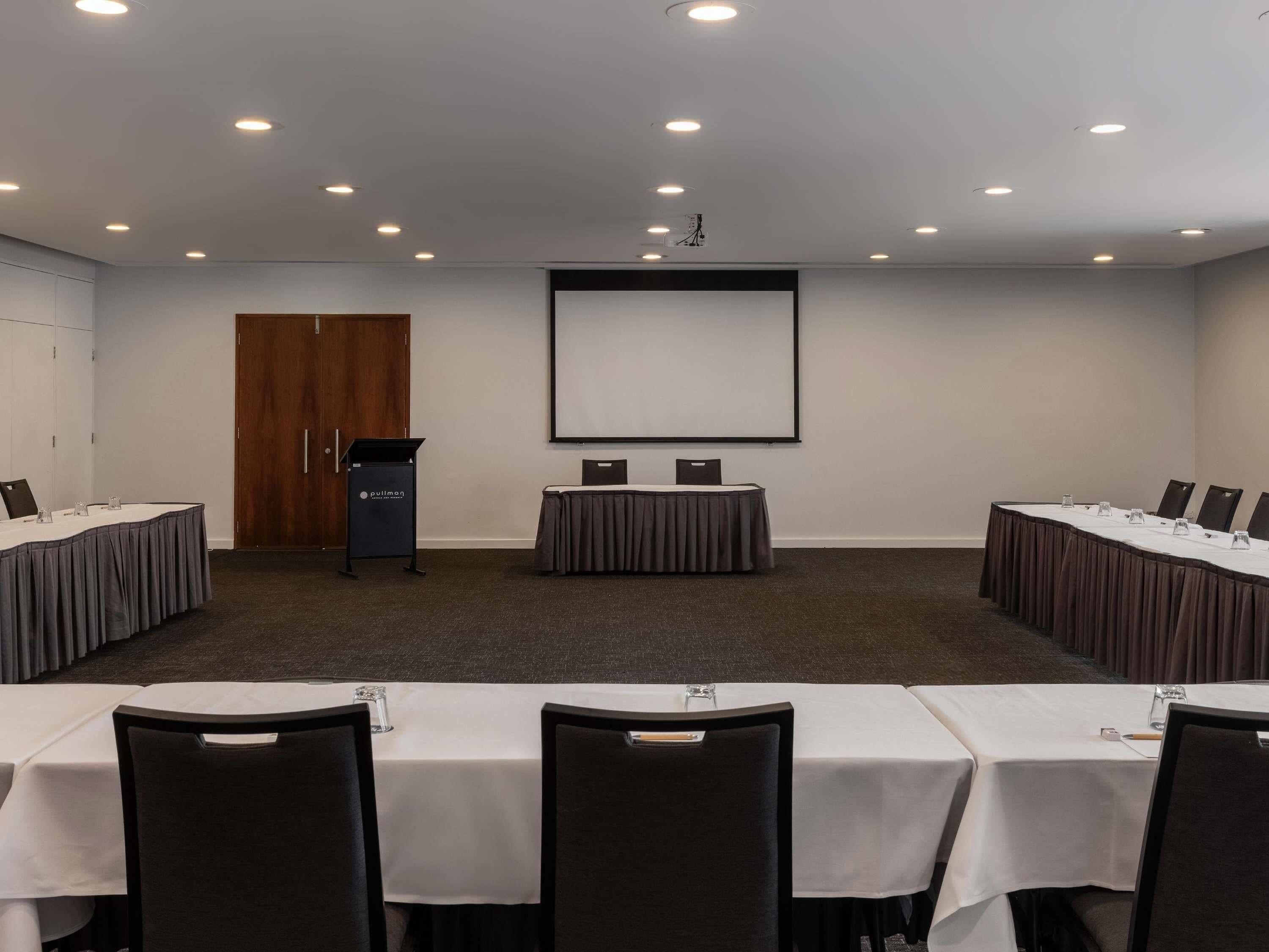 Una sala conferenze con un grande schermo, diversi tavoli con sedie e un podio.