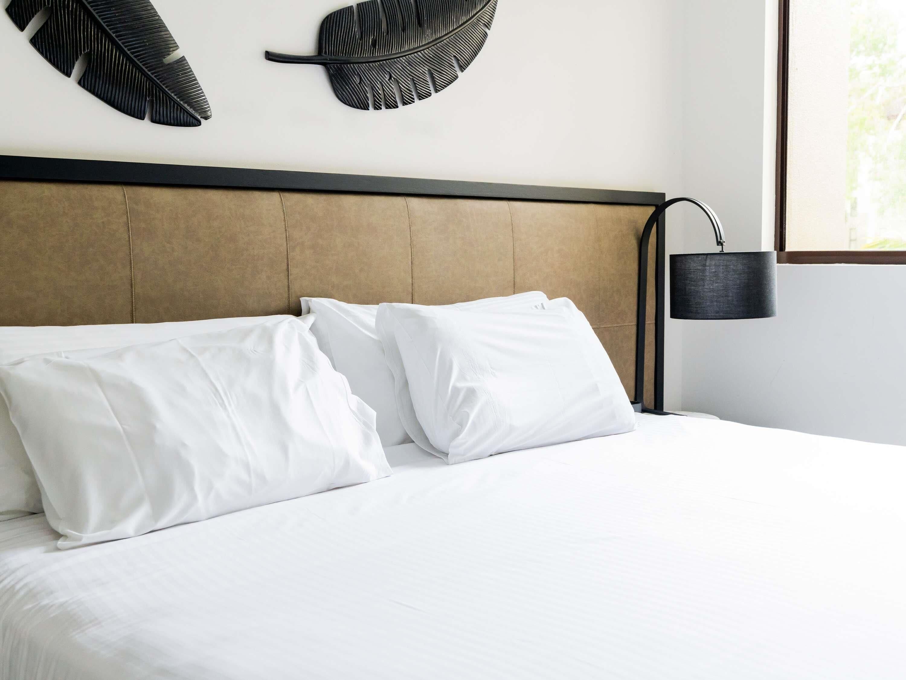 Un lit bien fait, avec du linge de lit blanc et des oreillers, une tête de lit marron et une lampe noire fixée au mur.
