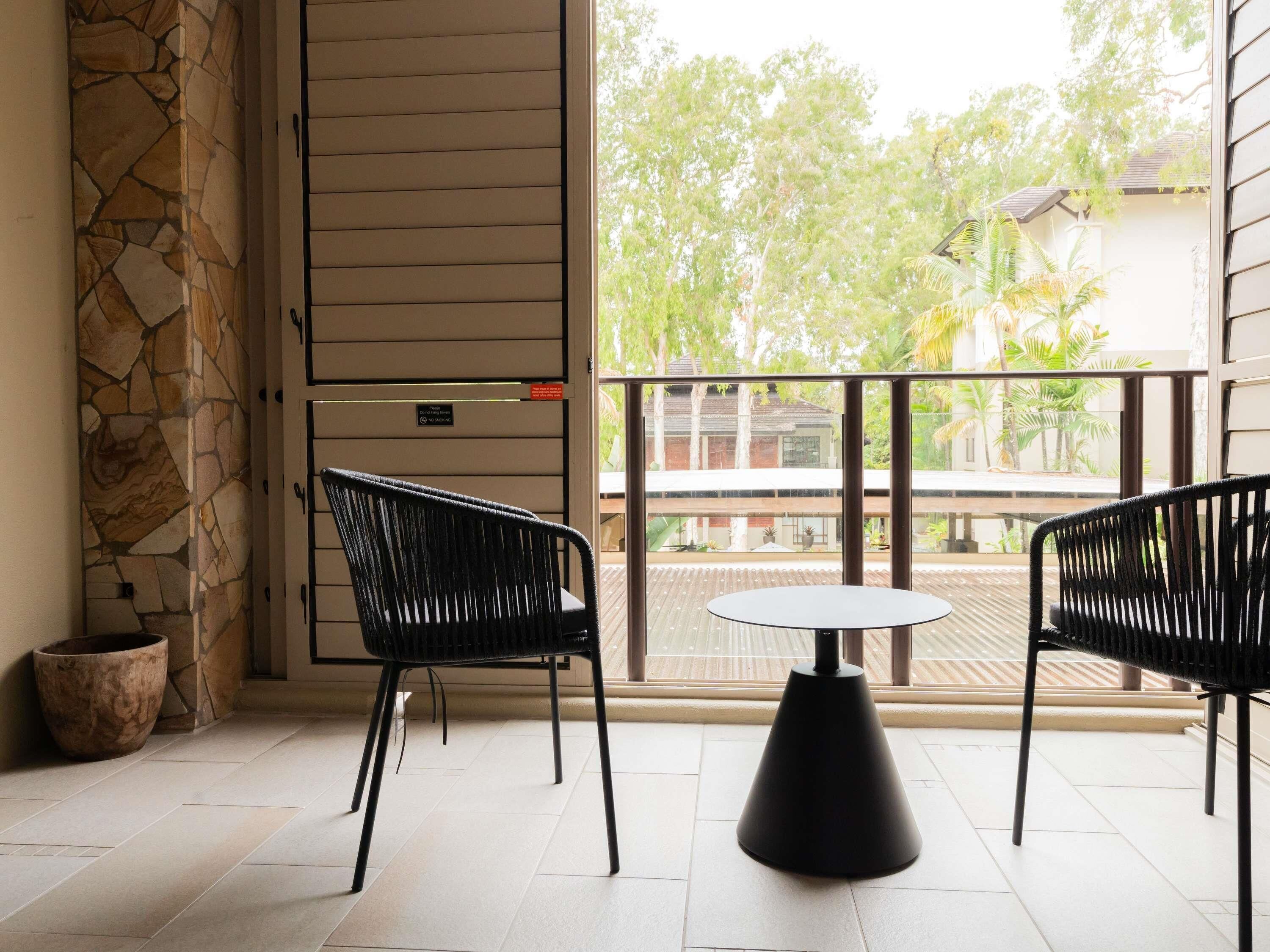 A balcony with two black chairs and a round table, a stone wall, and a view of trees and buildings.