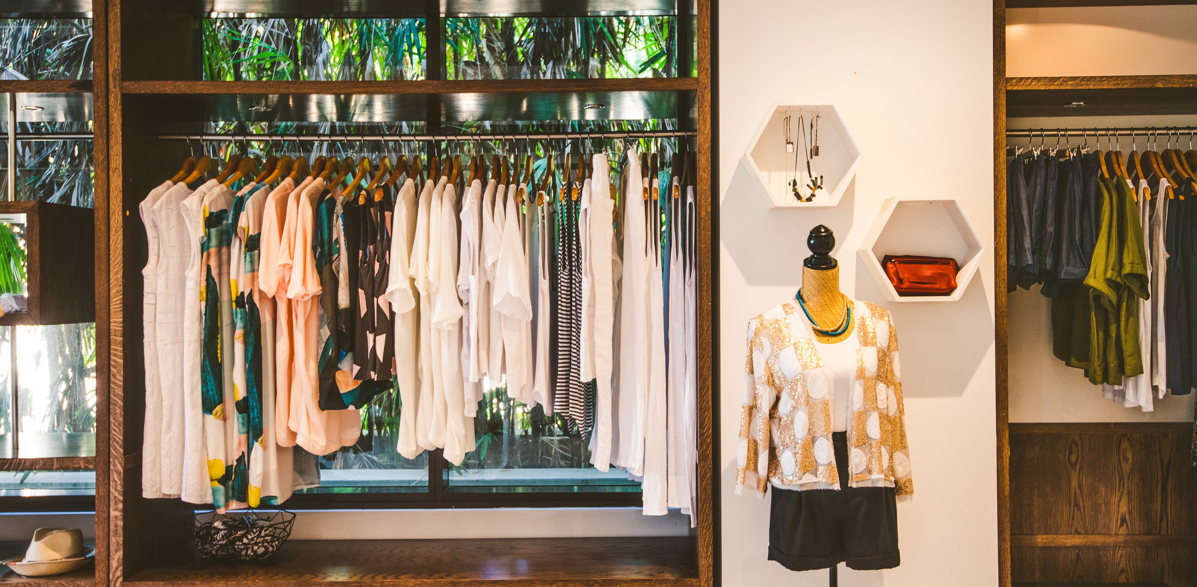 Un magasin de vêtements avec des portants remplis de vêtements, un mannequin et une vitrine ornée de plantes à l’extérieur.