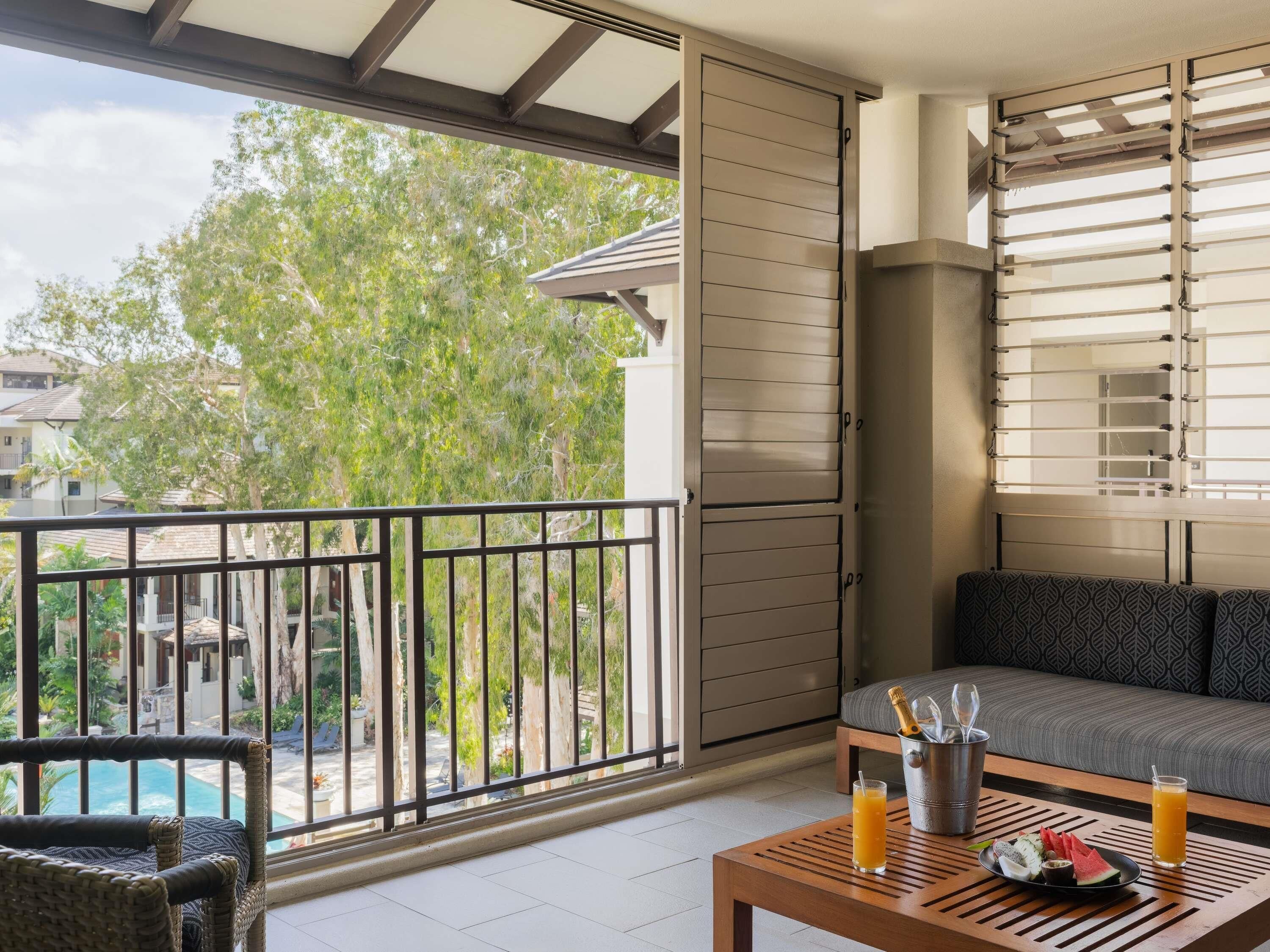 A balcony with a view of a pool and trees, a sofa, a table with drinks and snacks, and a sliding door.