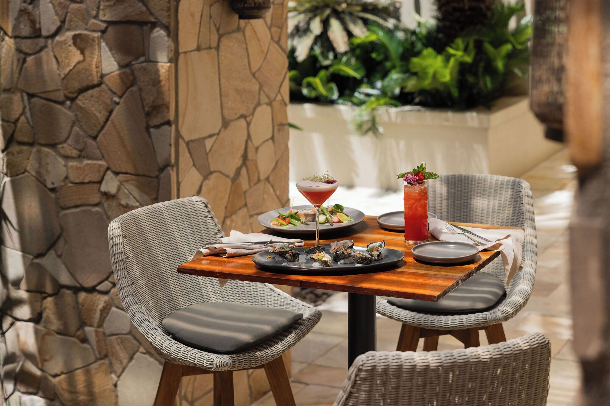 Una mesa de comedor con sillas de mimbre, una pared de piedra y una planta tropical al fondo.