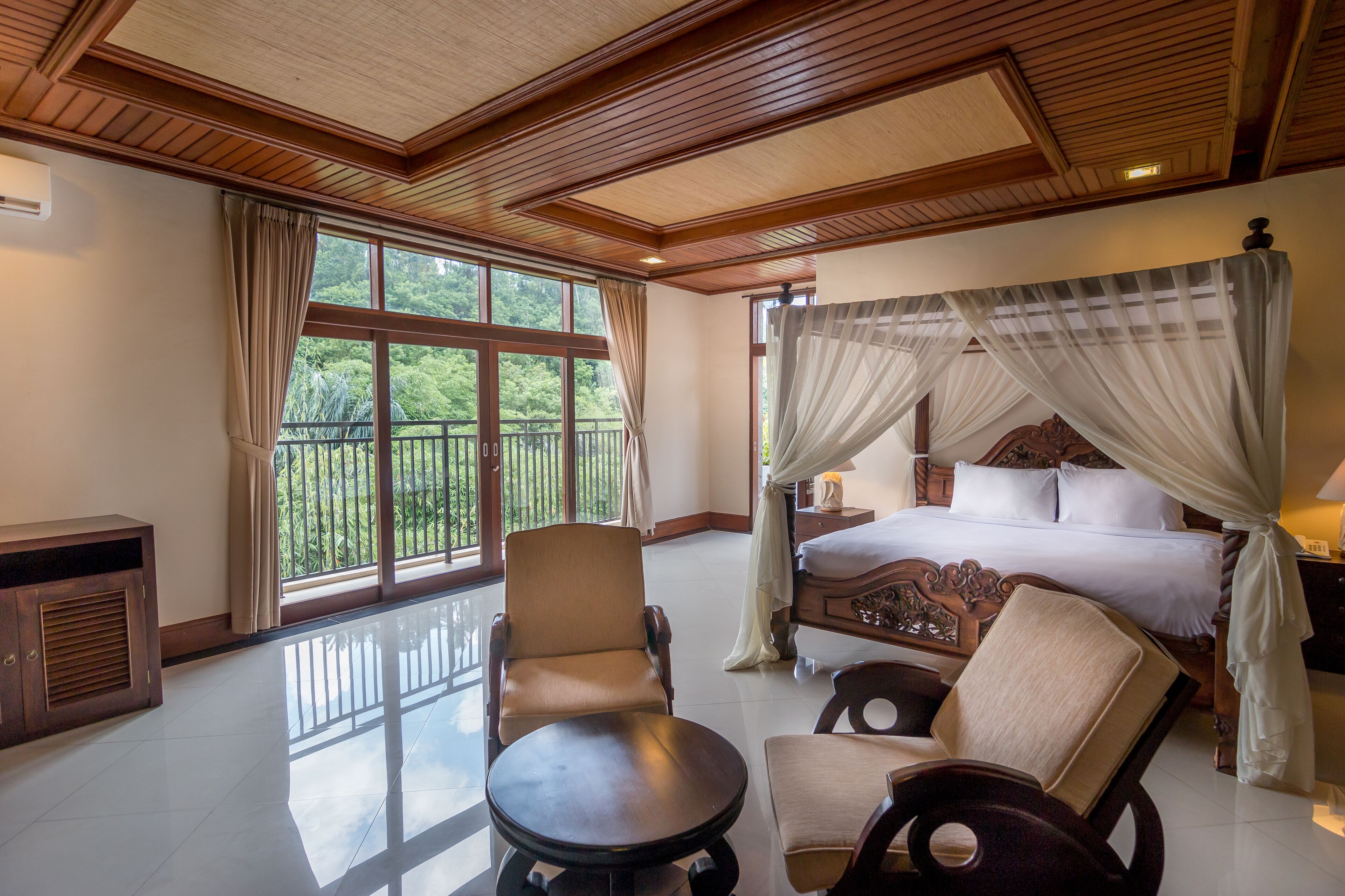 A spacious bedroom with a large bed, a sitting area with chairs, a small table, and a balcony with a view of greenery.
