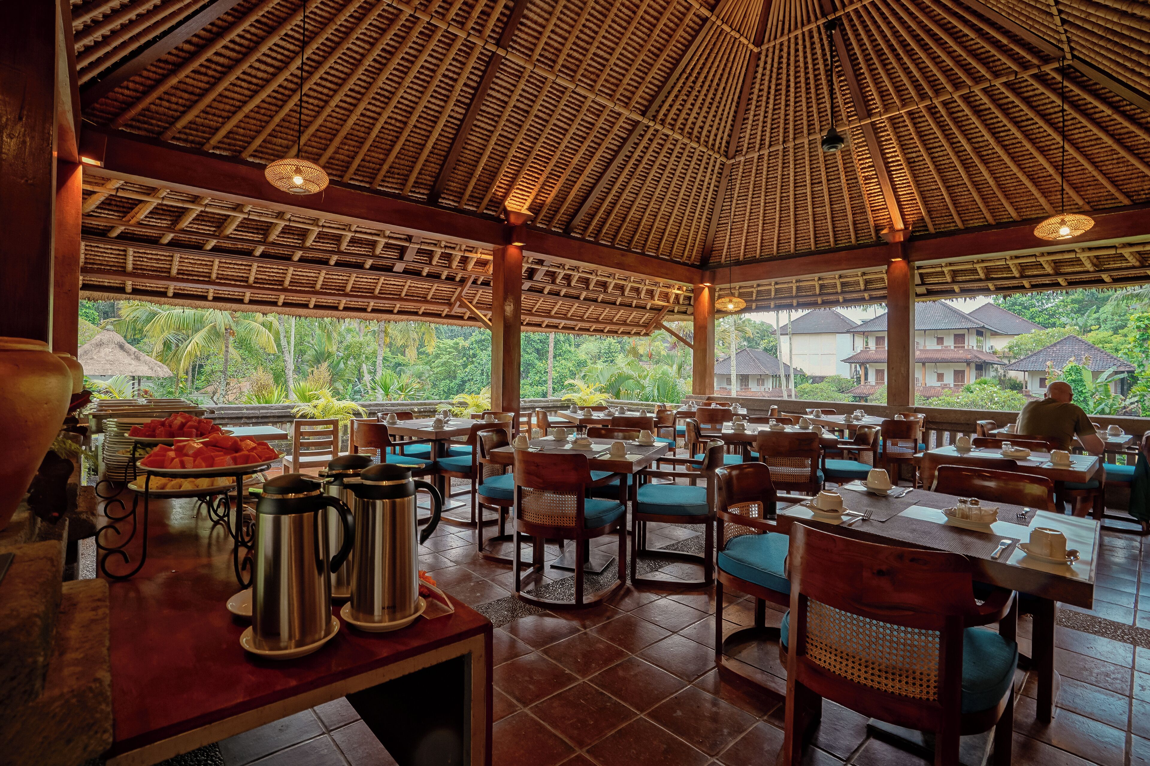 A restaurant with a thatched roof, wooden tables, and chairs, with a view of greenery outside.