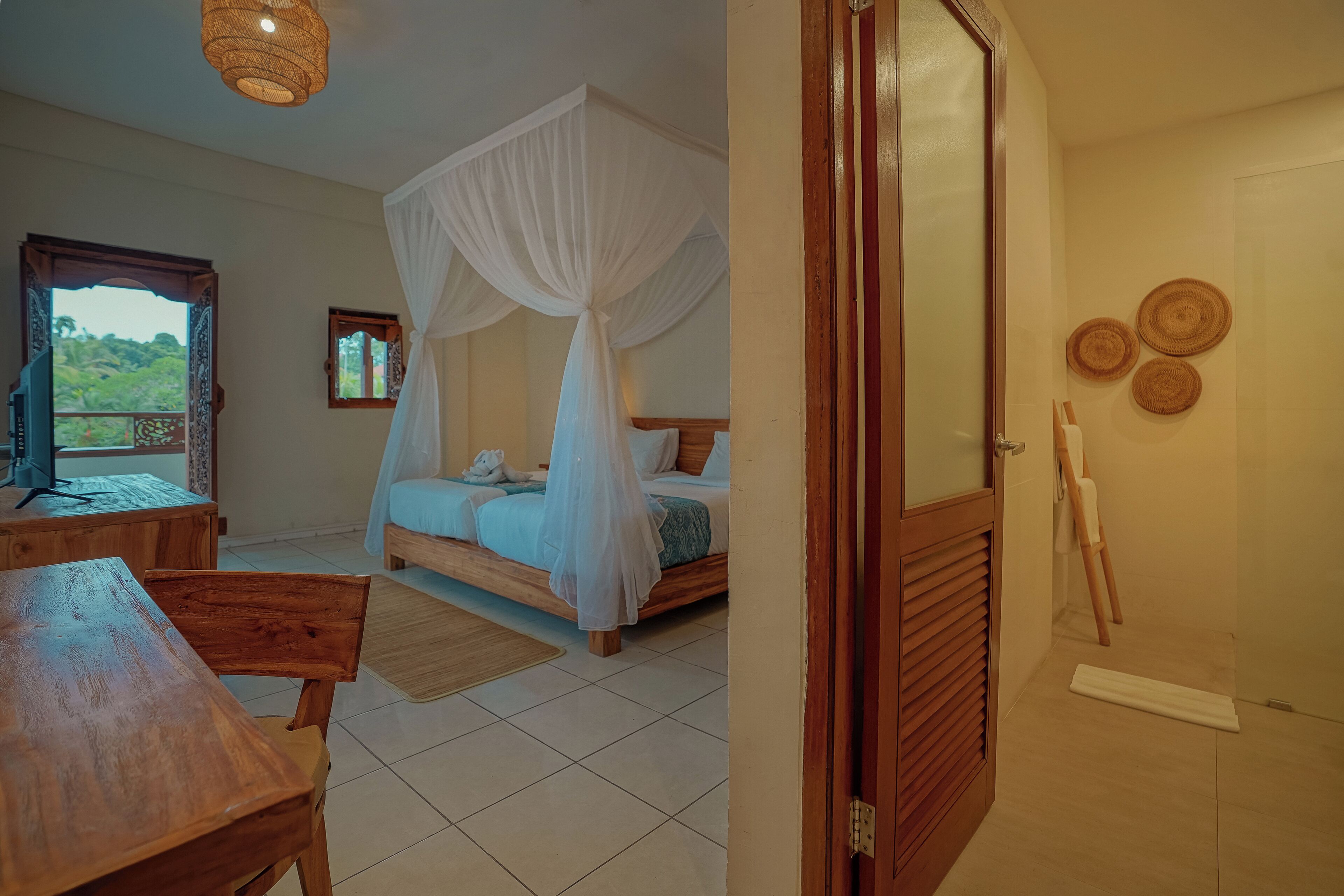 A bedroom with a bed under a canopy, a wooden table, and a view of greenery outside.