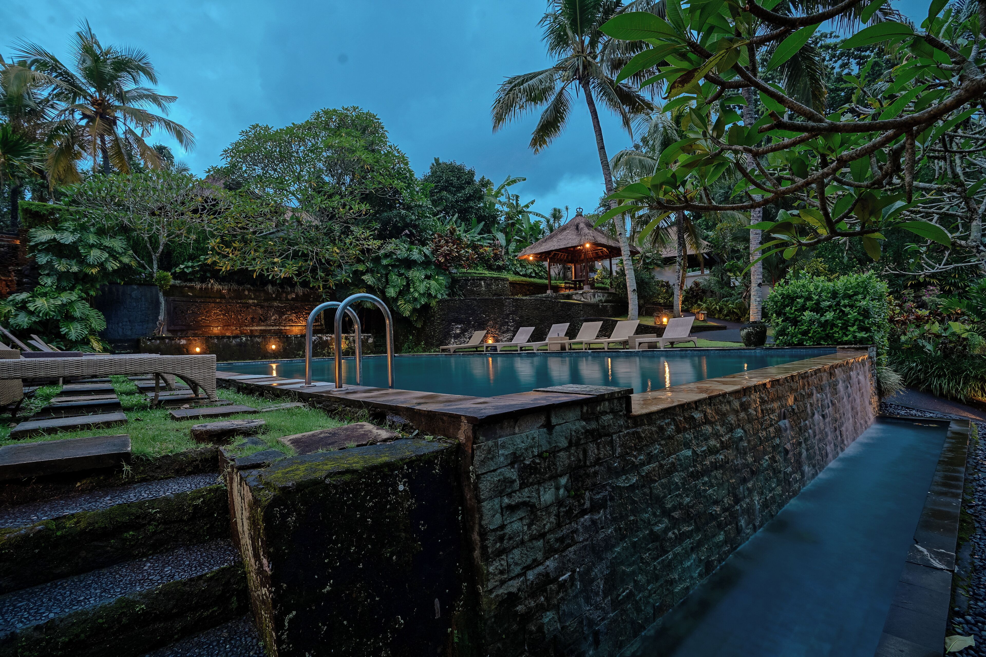 A serene outdoor pool area with lounge chairs and a thatched-roof structure, surrounded by lush greenery.
