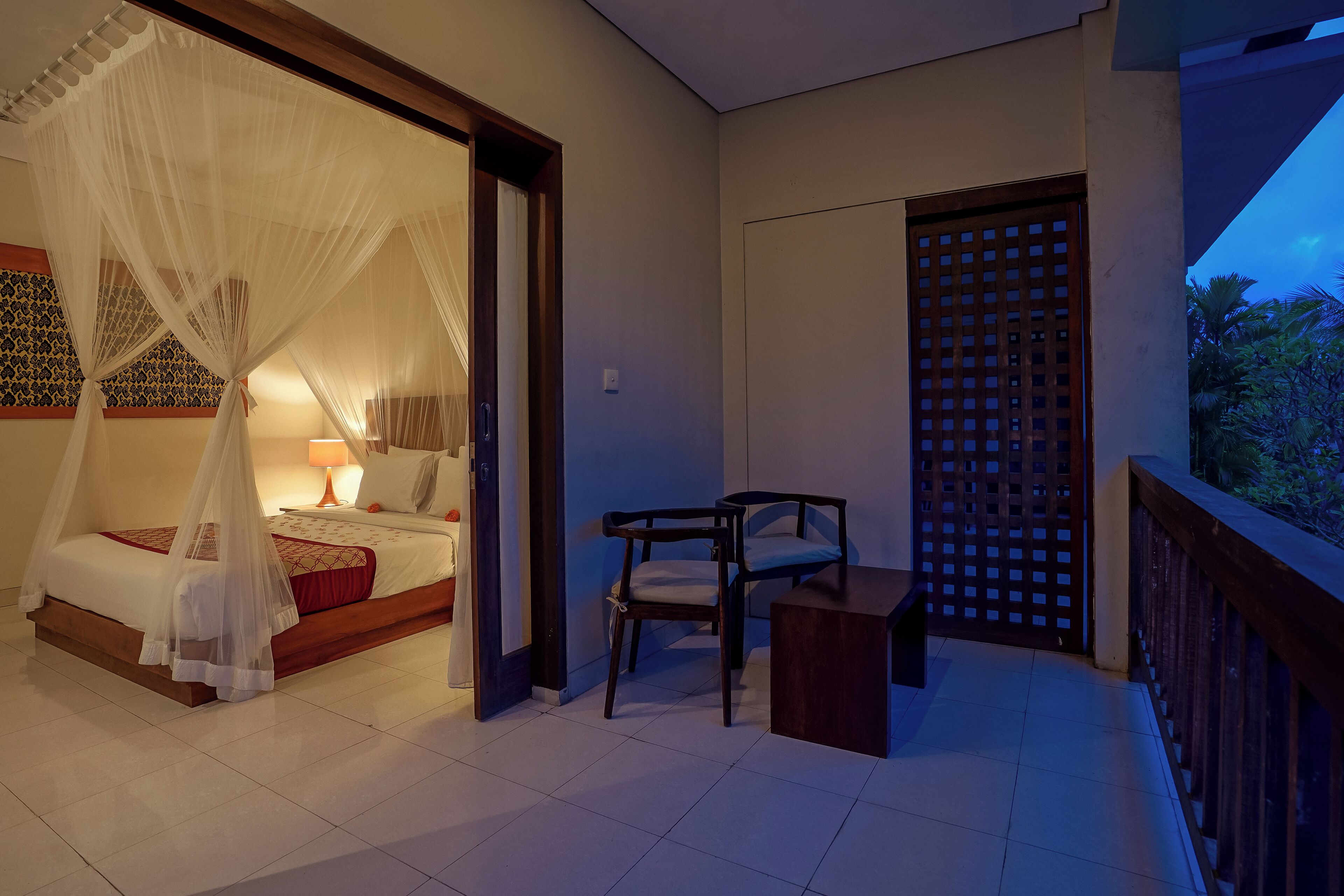 Ein Schlafzimmer mit Bett, Nachttisch und Stuhl, ein Balkon mit Holzbalkon und Blick auf Grünfläche.