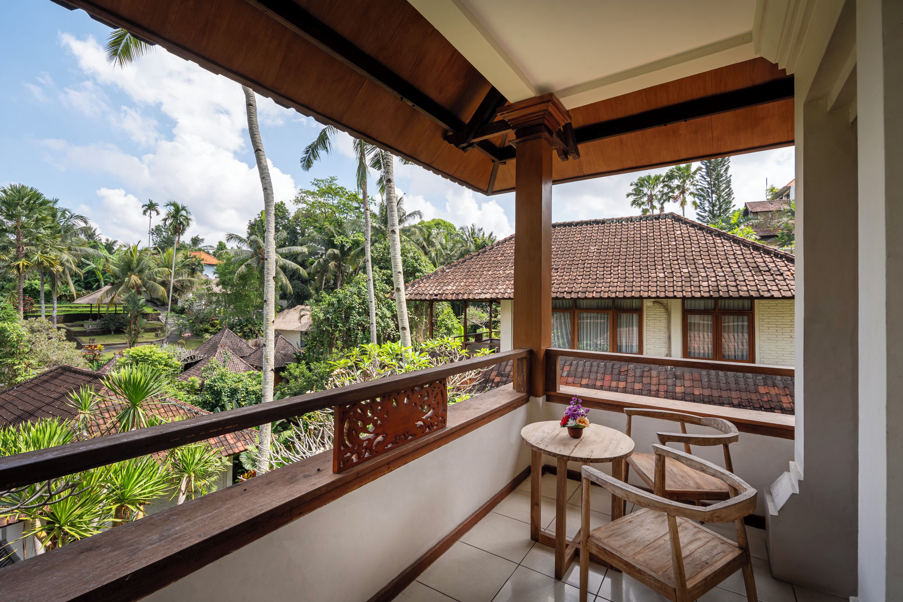 A balcony with wooden furniture, a view of a residential area with tiled roofs, and lush greenery.