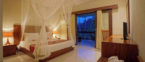 A bedroom with a four-poster bed, a wooden desk, a TV, and a balcony with a view of palm trees.