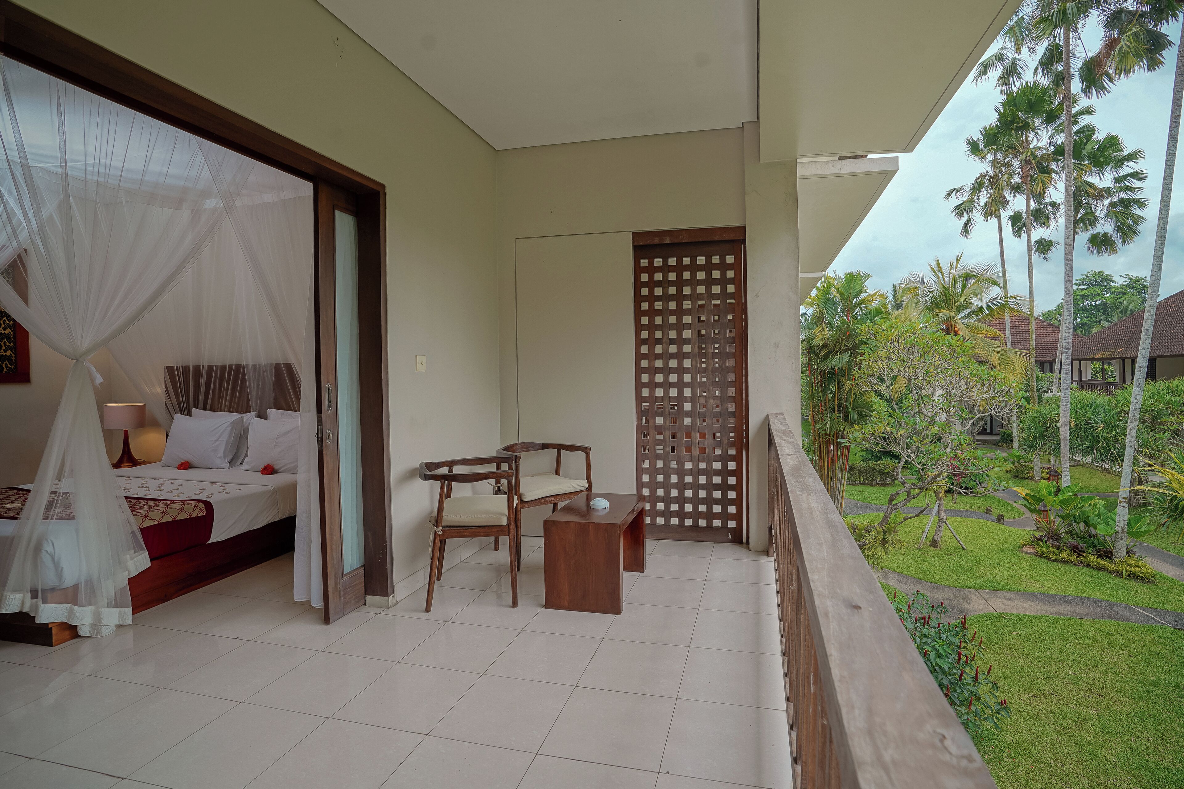 A bedroom with a bed, bedside table, and chair, a balcony with a view of the garden and palm trees.