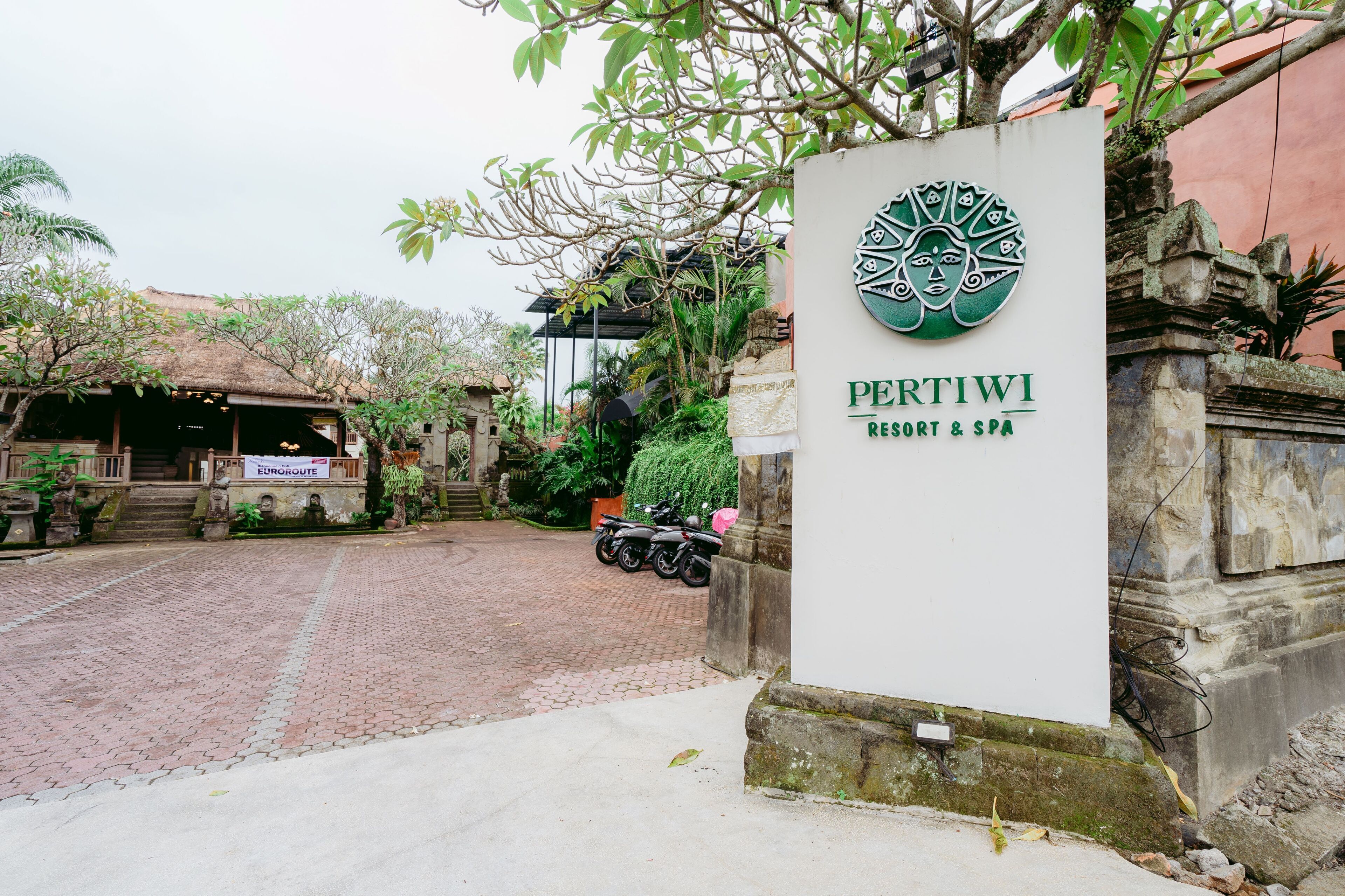 A sign for PertiwI Resort & Spa with a traditional design, a paved entrance, and a parked motorcycle.