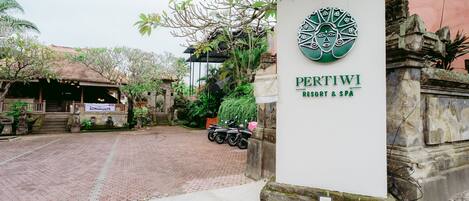A sign for PertiwI Resort & Spa with a traditional design, a paved entrance, and a parked motorcycle.