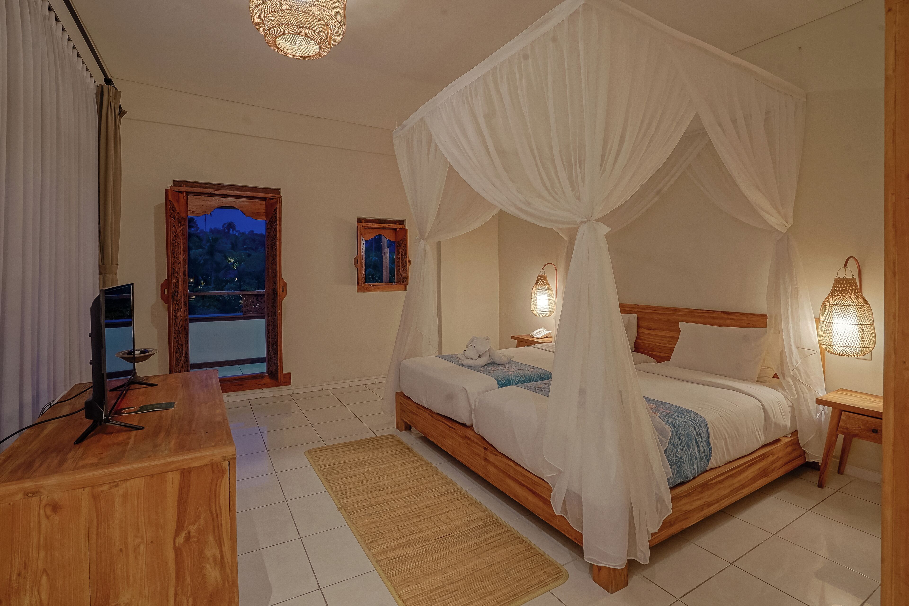Un dormitorio con cama de madera, dosel blanco, lámparas de noche, un escritorio con computadora y vistas al exterior.