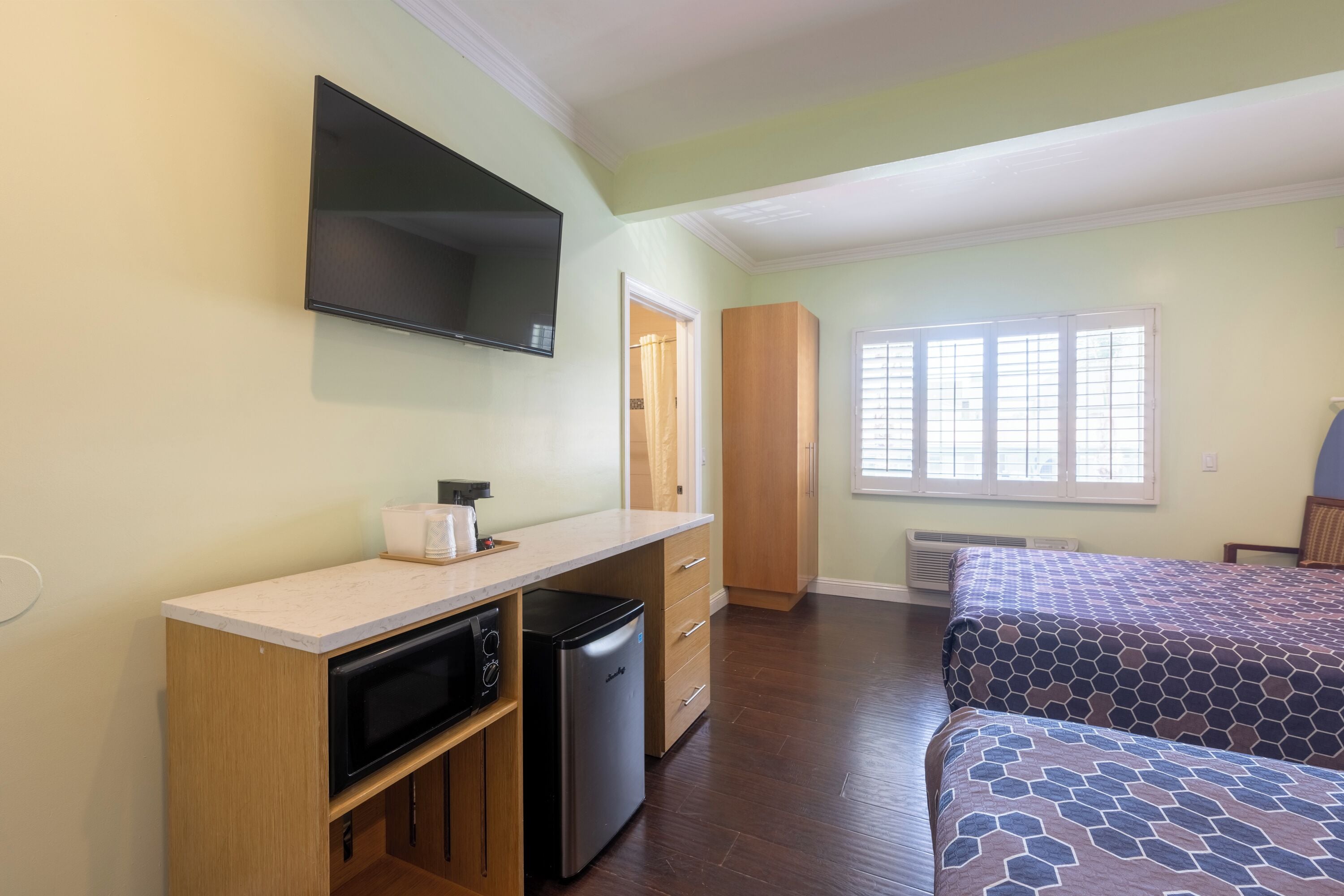 A compact room with a kitchenette, a bed with patterned bedding, and a window with blinds.
