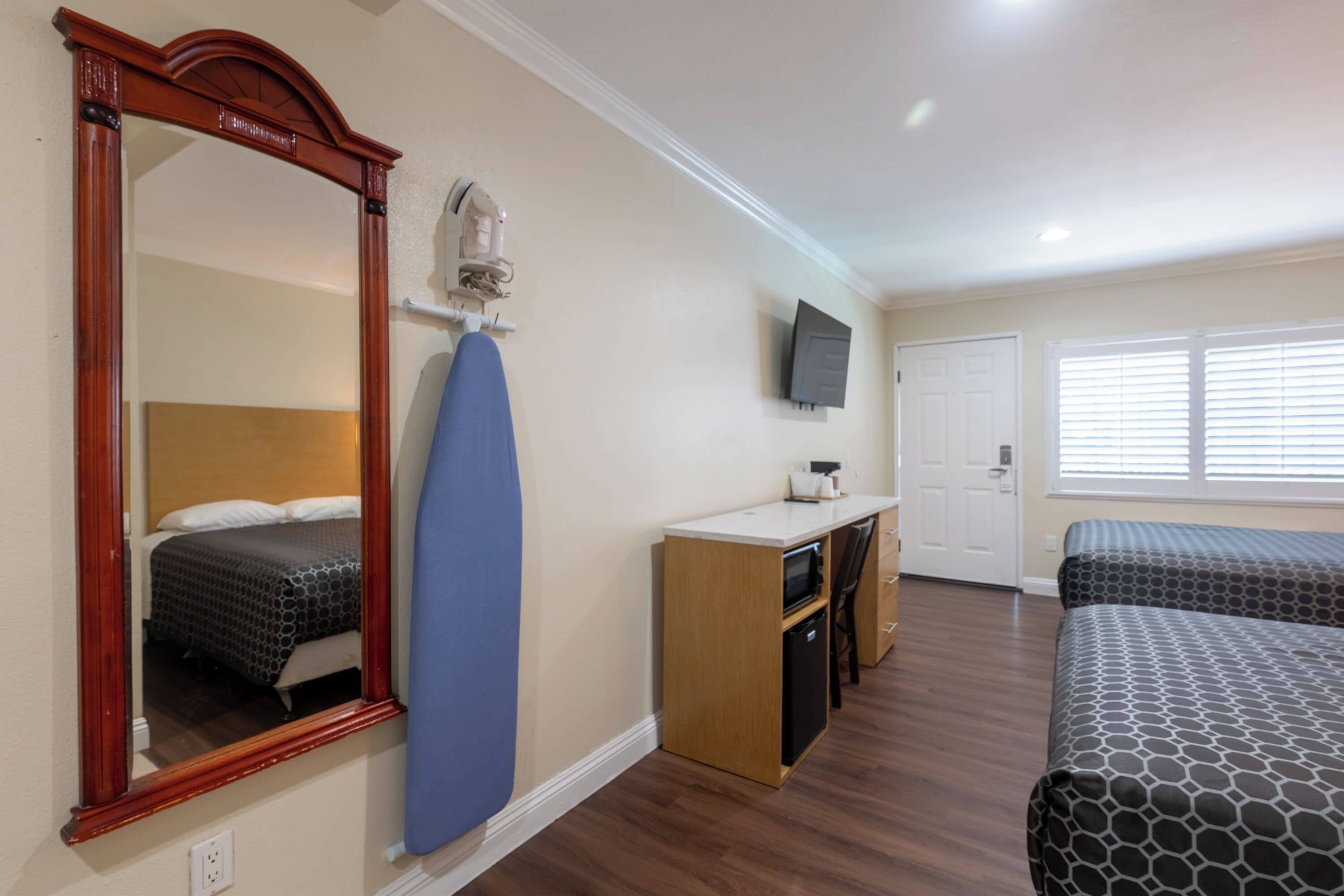 A hotel room with a wooden floor, a large mirror, a desk with a chair, a television mounted on the wall, and two beds with patterned bedding.