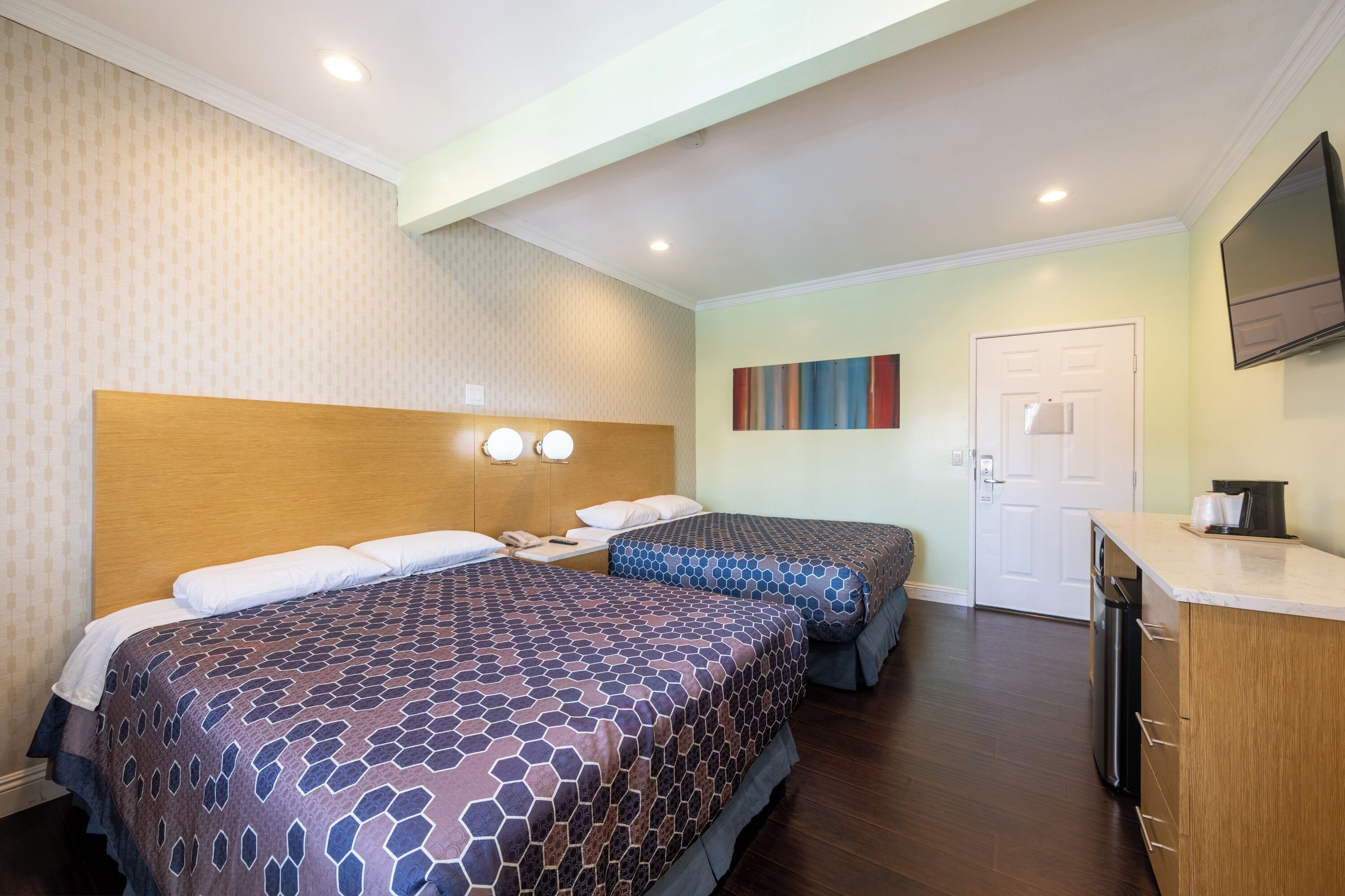 A hotel room with two beds, a wooden headboard, a flat-screen TV, a small kitchenette, and a door leading to the bathroom.