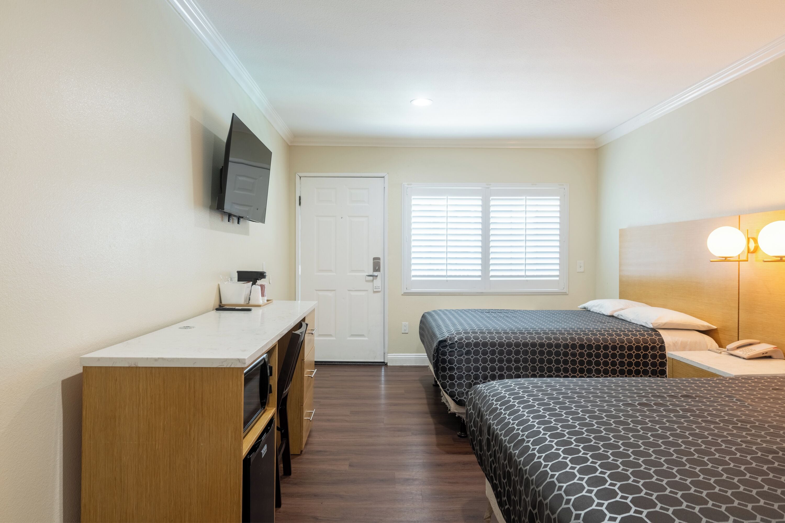 A hotel room with two beds, a desk, a TV mounted on the wall, and a window with blinds.