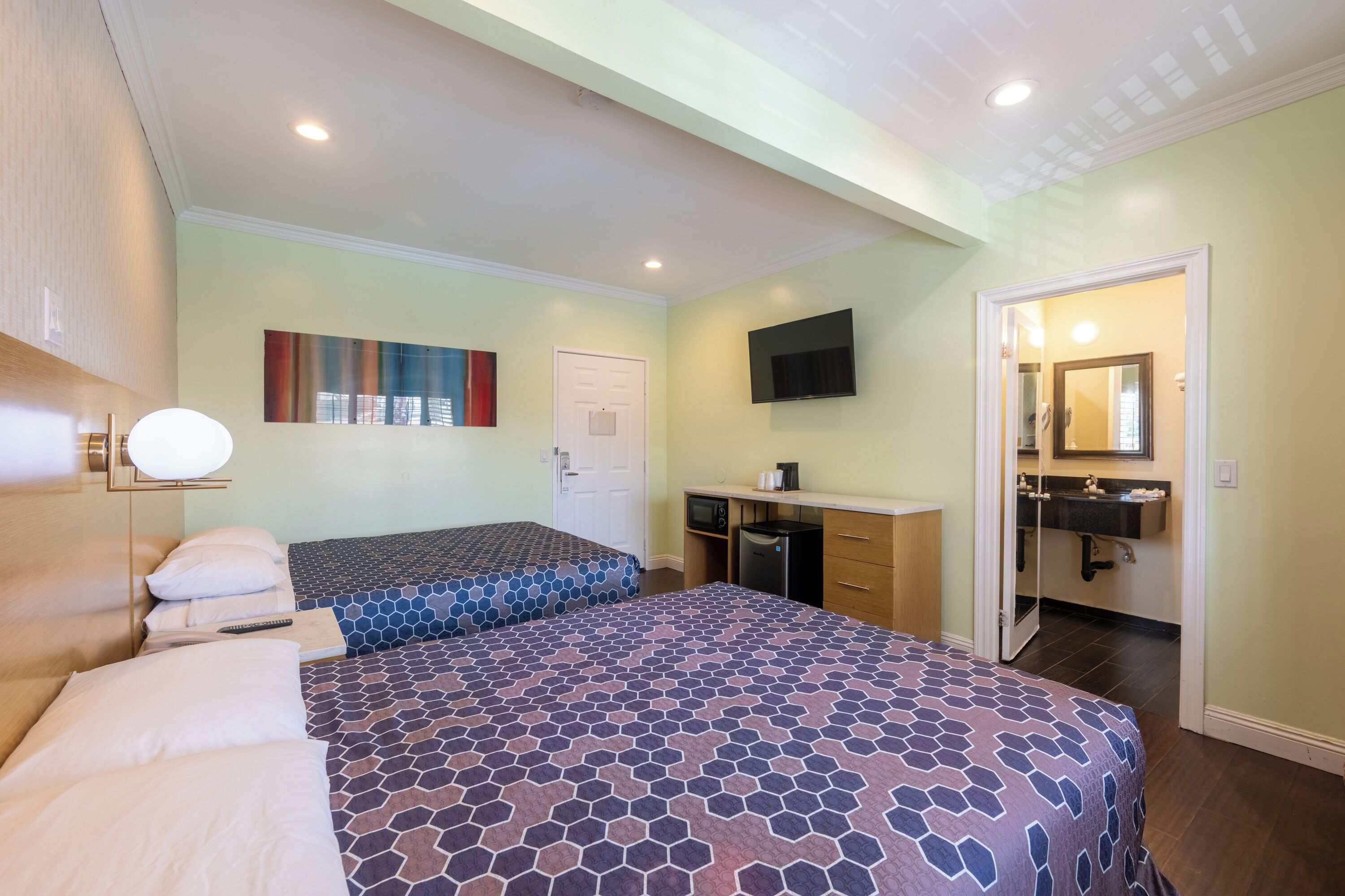 A hotel room with two beds, a desk with a computer, a TV, and a bathroom with a sink and mirror.