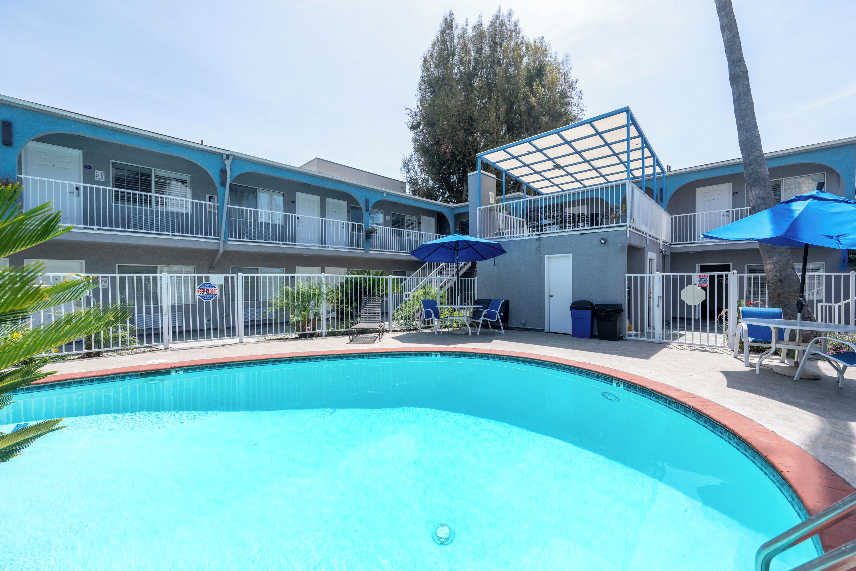 A swimming pool with lounge chairs and umbrellas, surrounded by a fenced area with a two-story building in the background.