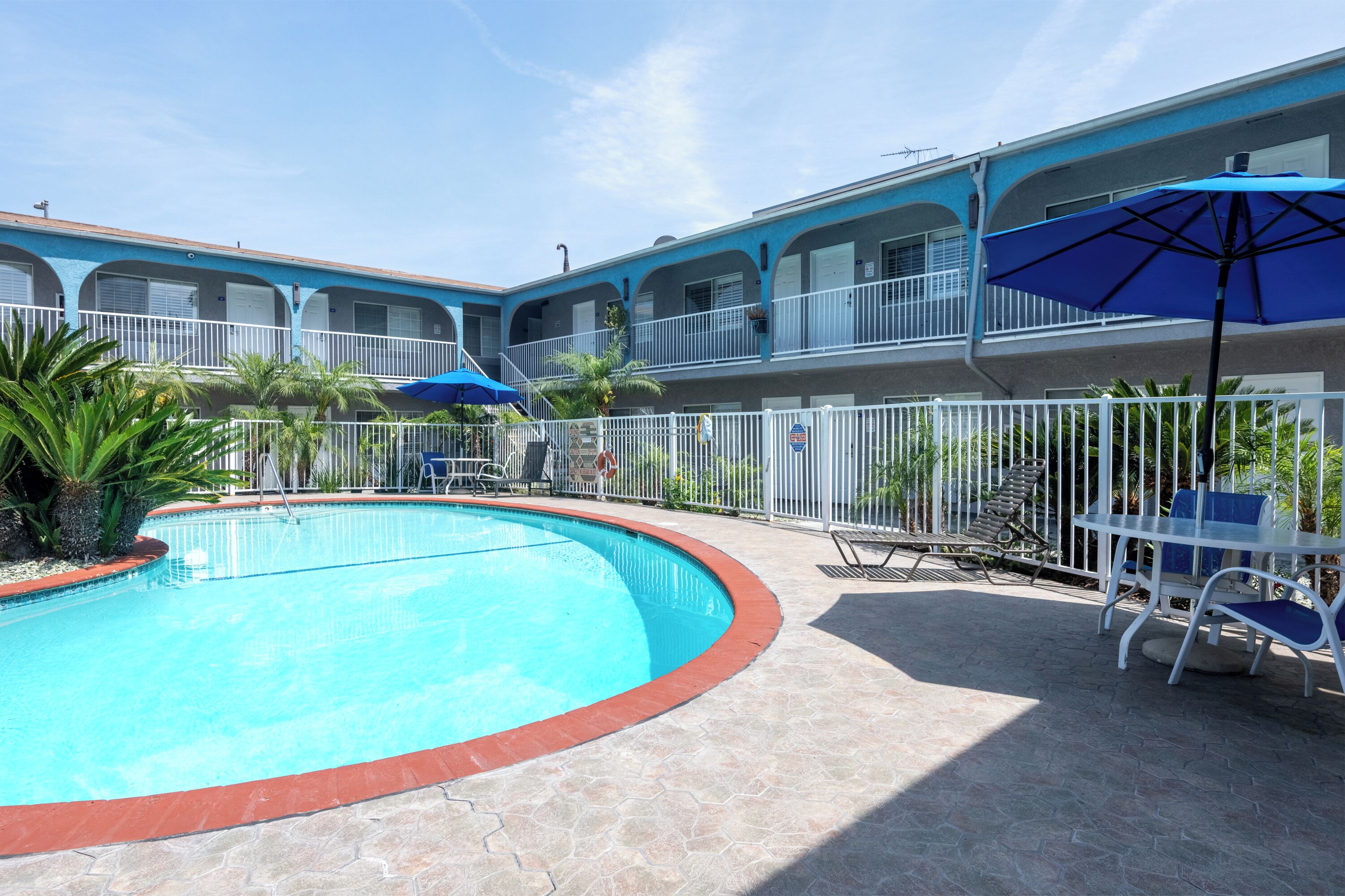 A swimming pool with a red border, surrounded by a white fence and palm trees. There are lounge chairs and umbrellas.
