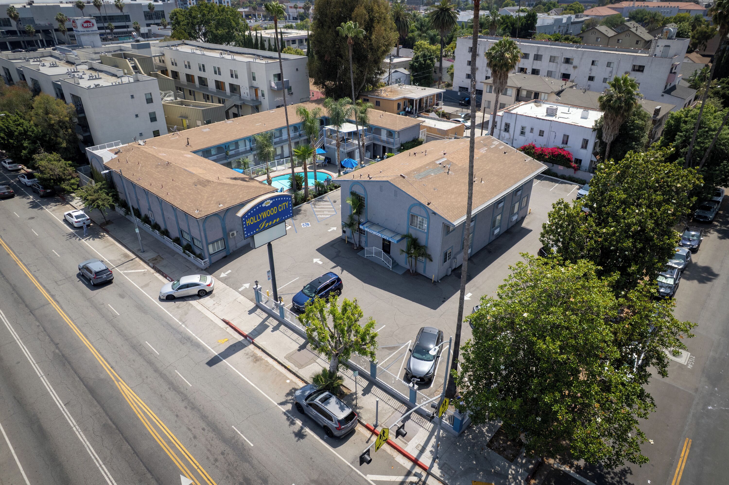 An aerial view of a multi-story building with a pool, surrounded by palm trees and parked cars.