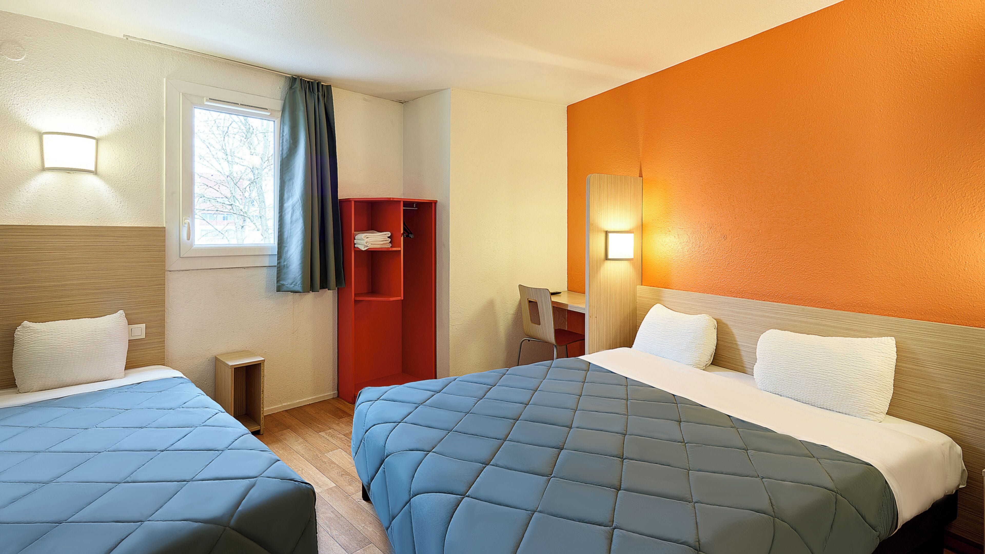 A hotel room with two beds, a desk, a chair, a wardrobe, and a window with curtains.