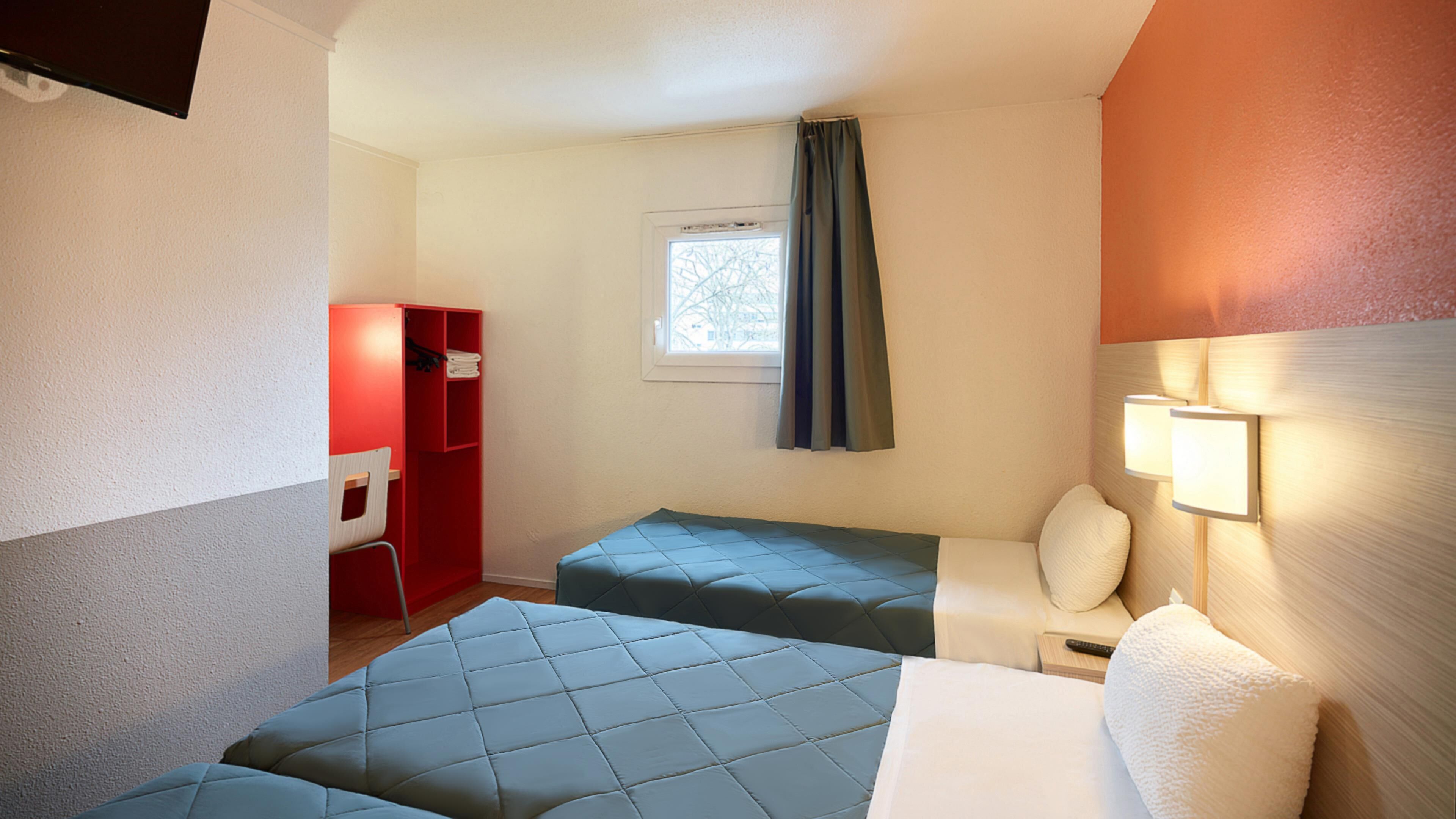 A hotel room with two beds, a red wardrobe, a window with curtains, and a bedside lamp.