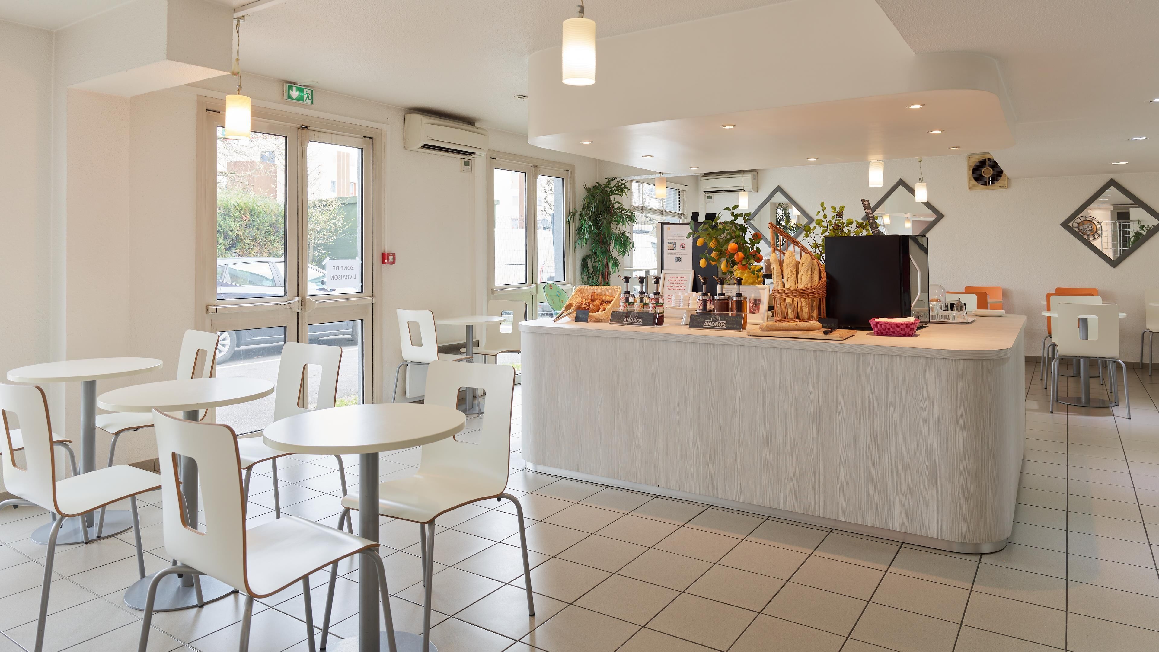 A clean, modern cafe interior with white furniture, a central counter, and a view of the street outside.